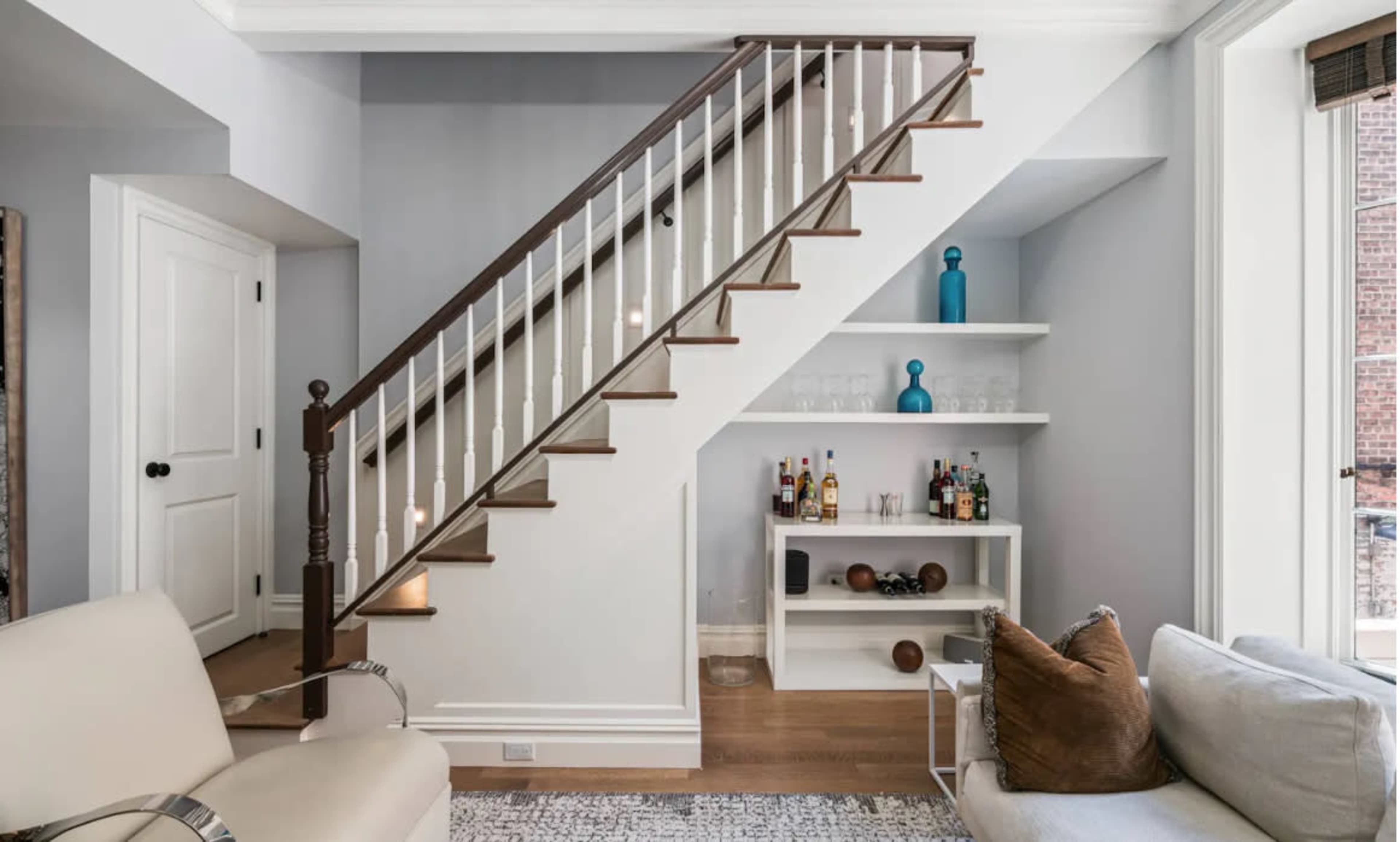 A staircase with white railings ascends to a landing beside a small shelving unit and a seating area.