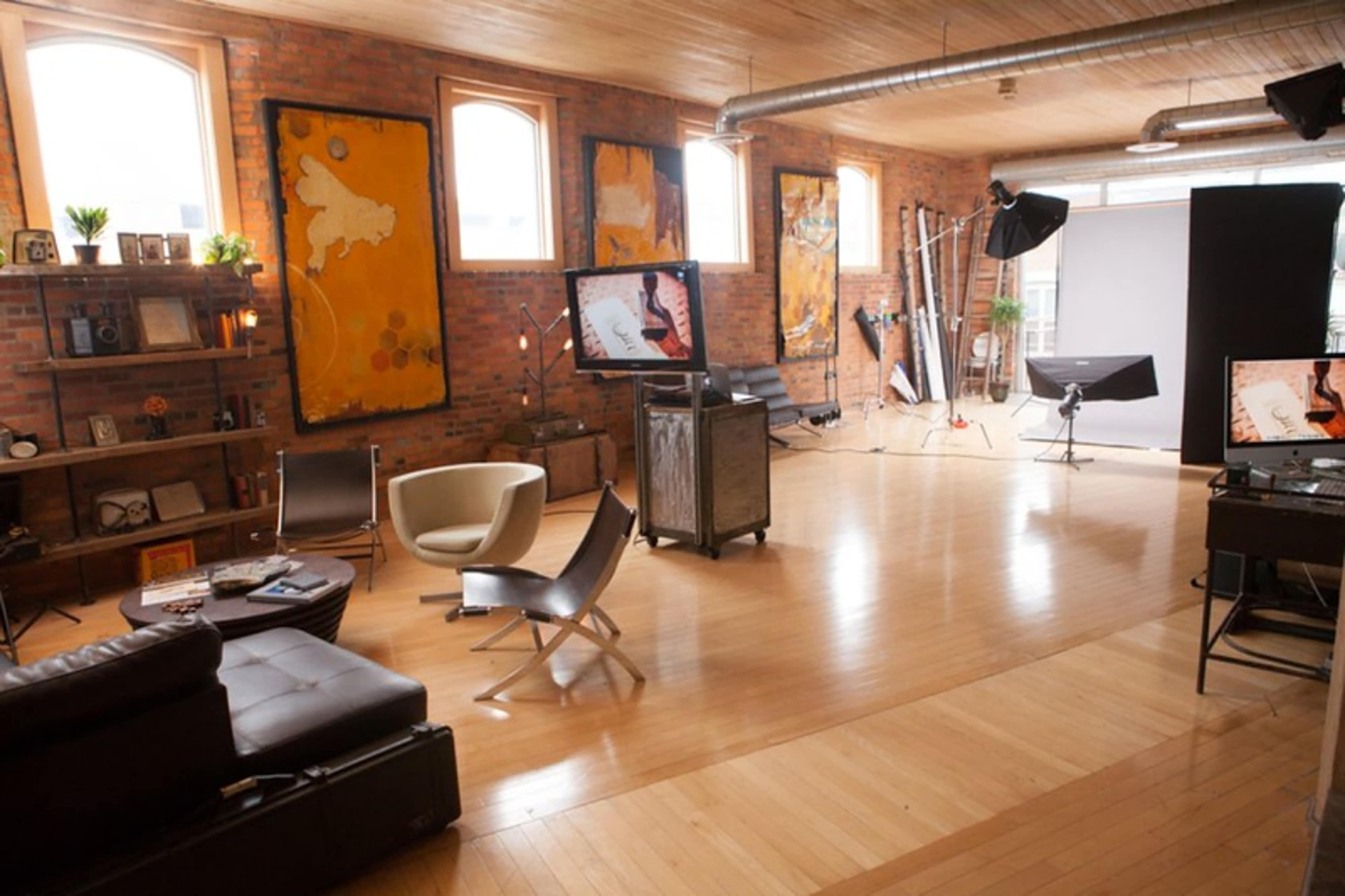 A spacious studio with wooden floors, large windows, and various pieces of furniture, including modern chairs and a leather couch, along with photography equipment set up in one corner.
