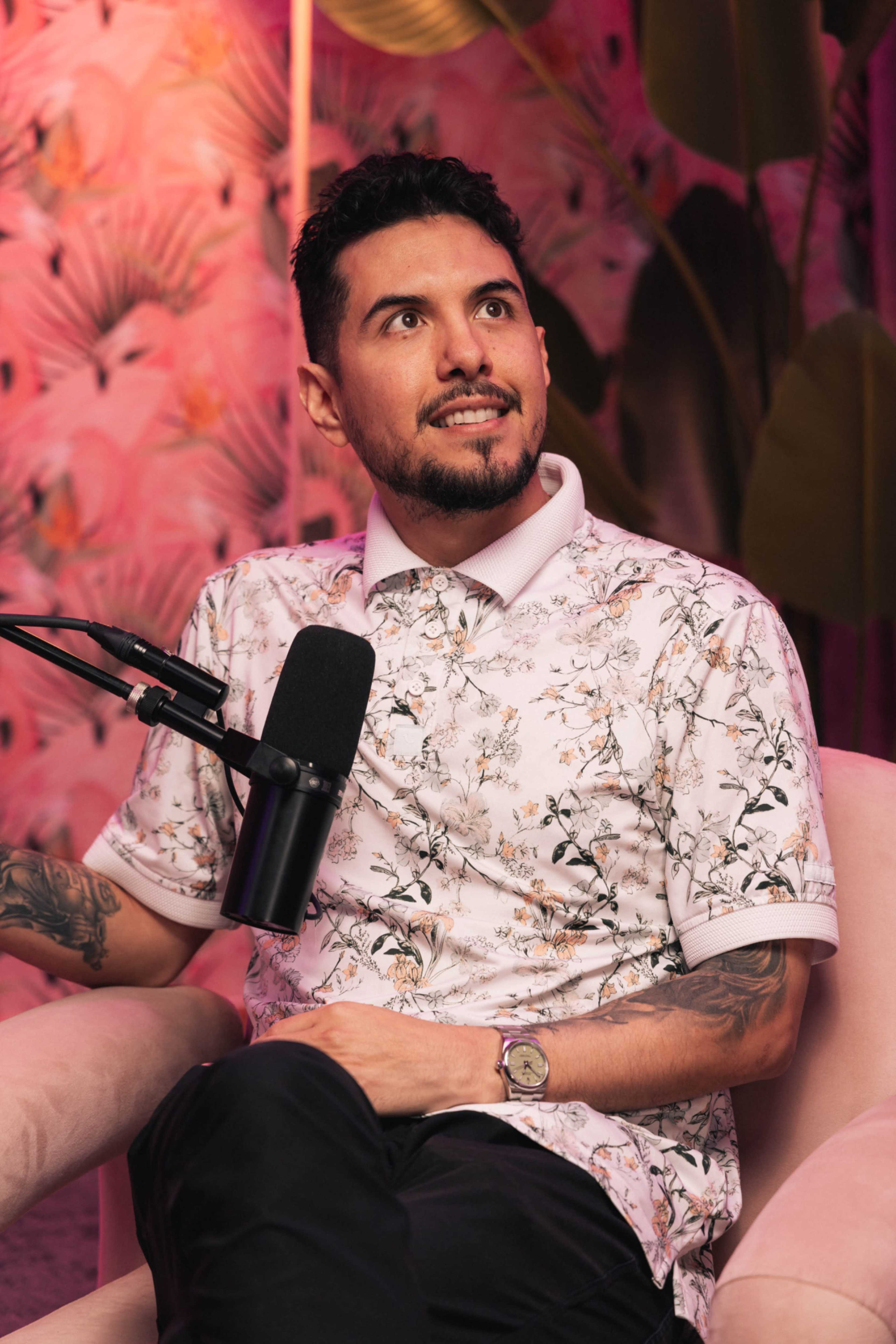 A person with tattoos is sitting in a chair, wearing a floral-patterned polo shirt, while speaking into a microphone in a brightly decorated room with pink and floral backgrounds.