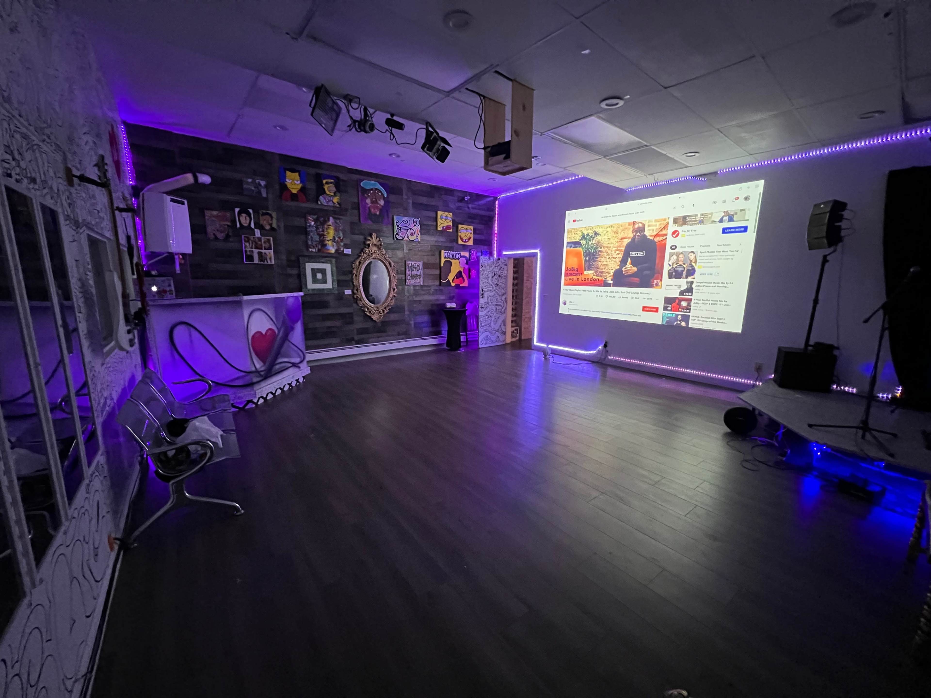 A modern studio space with a large projector screen displaying a video, surrounded by colorful artwork and illuminated by purple lights.