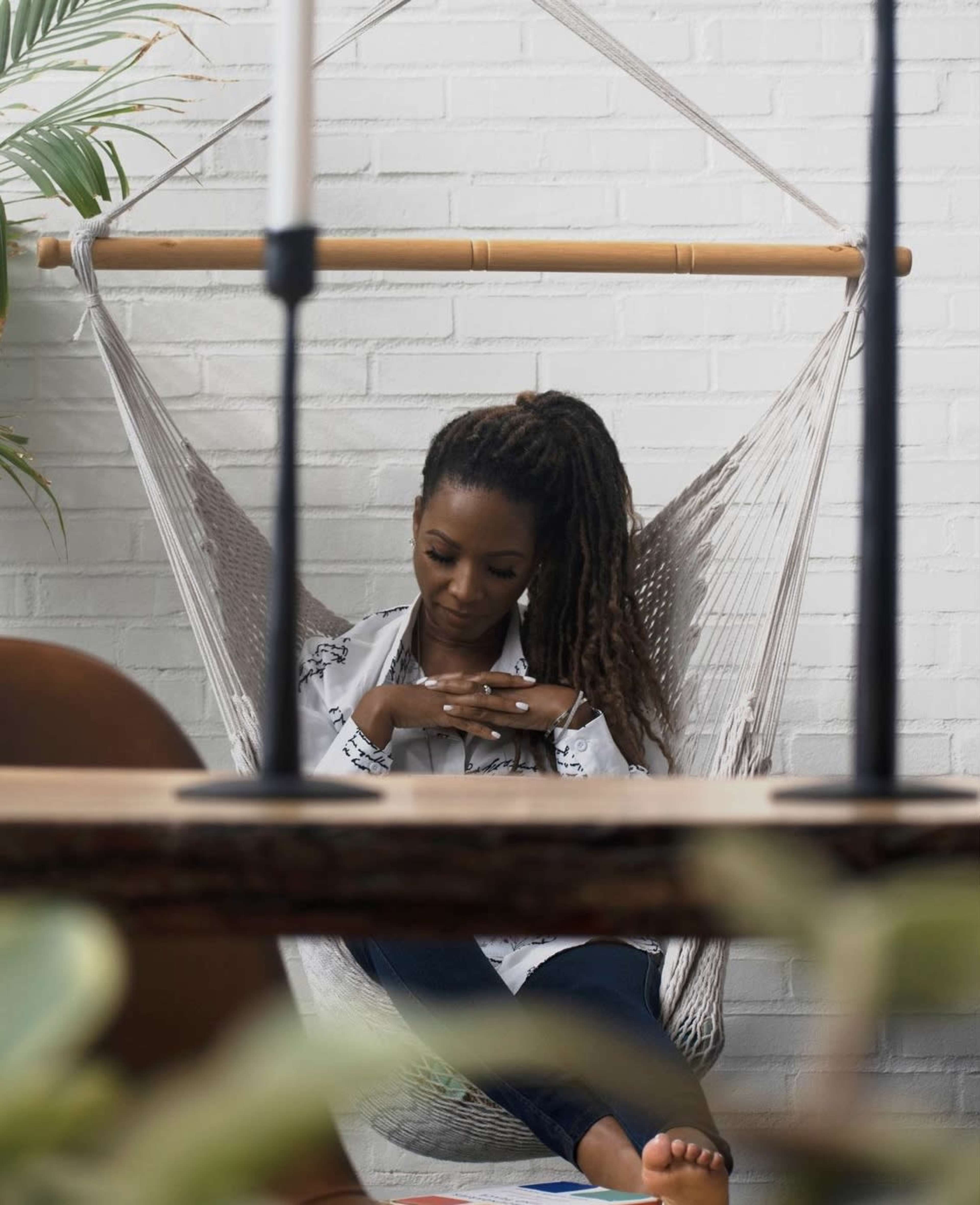 A woman sits in a hammock, focused on her phone, with a white brick wall and indoor plants in the background.