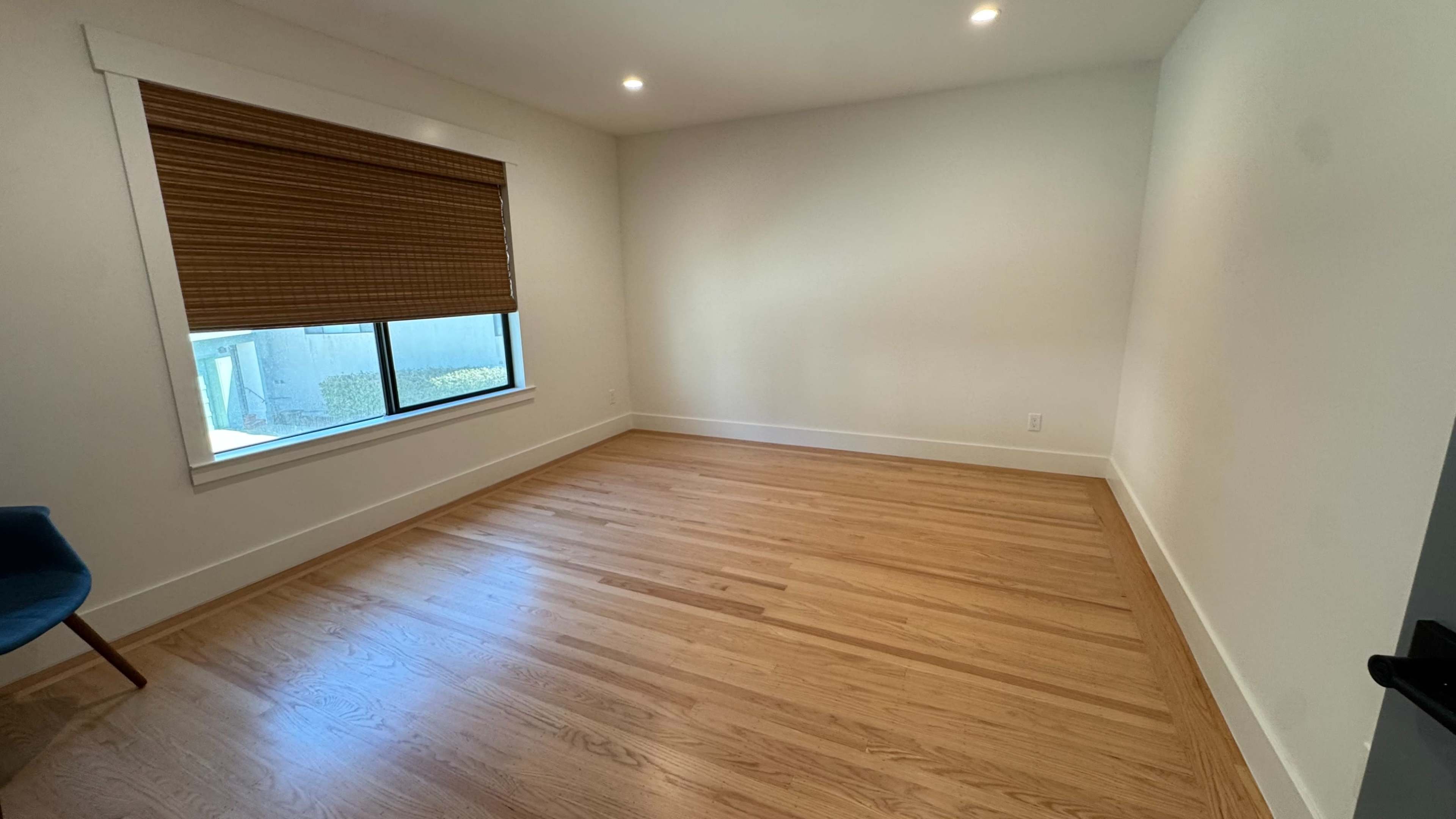 The image shows an empty room with hardwood floors, a single window covered by a wooden blind, and a blue chair in the corner.