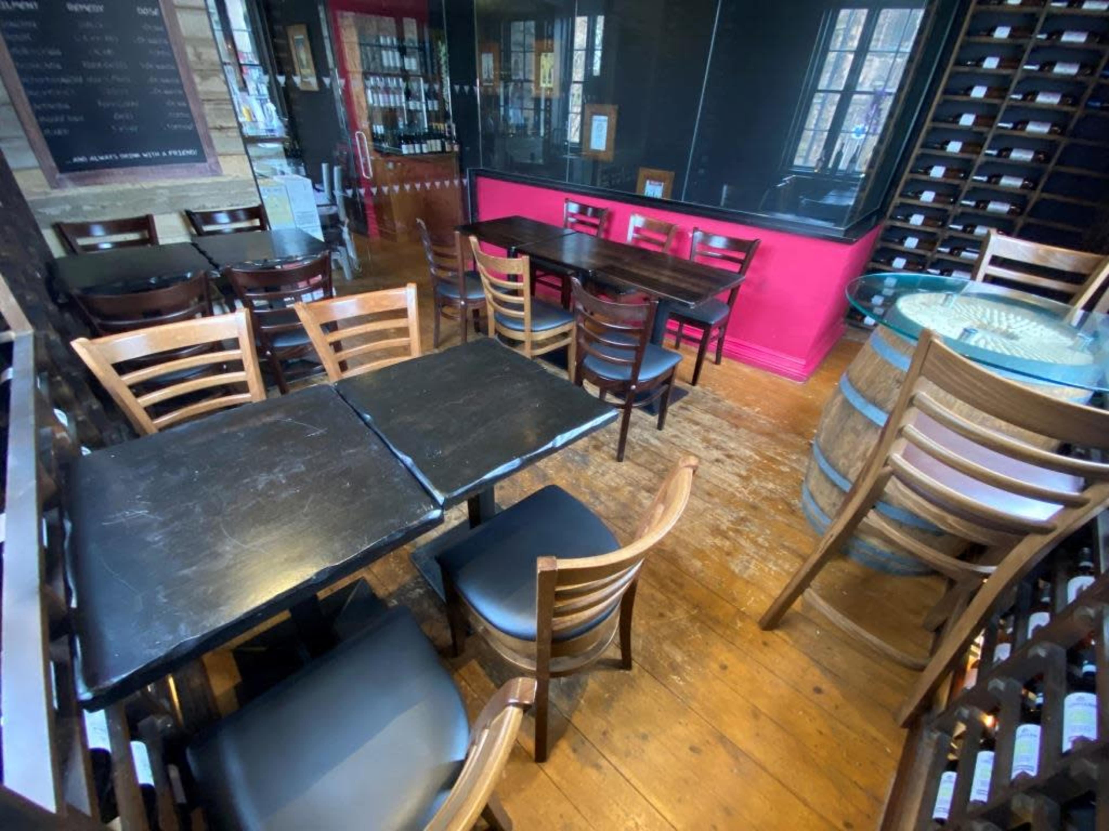 The image shows a cozy restaurant interior with wooden floors, several tables and chairs, and a wall lined with wine bottles.