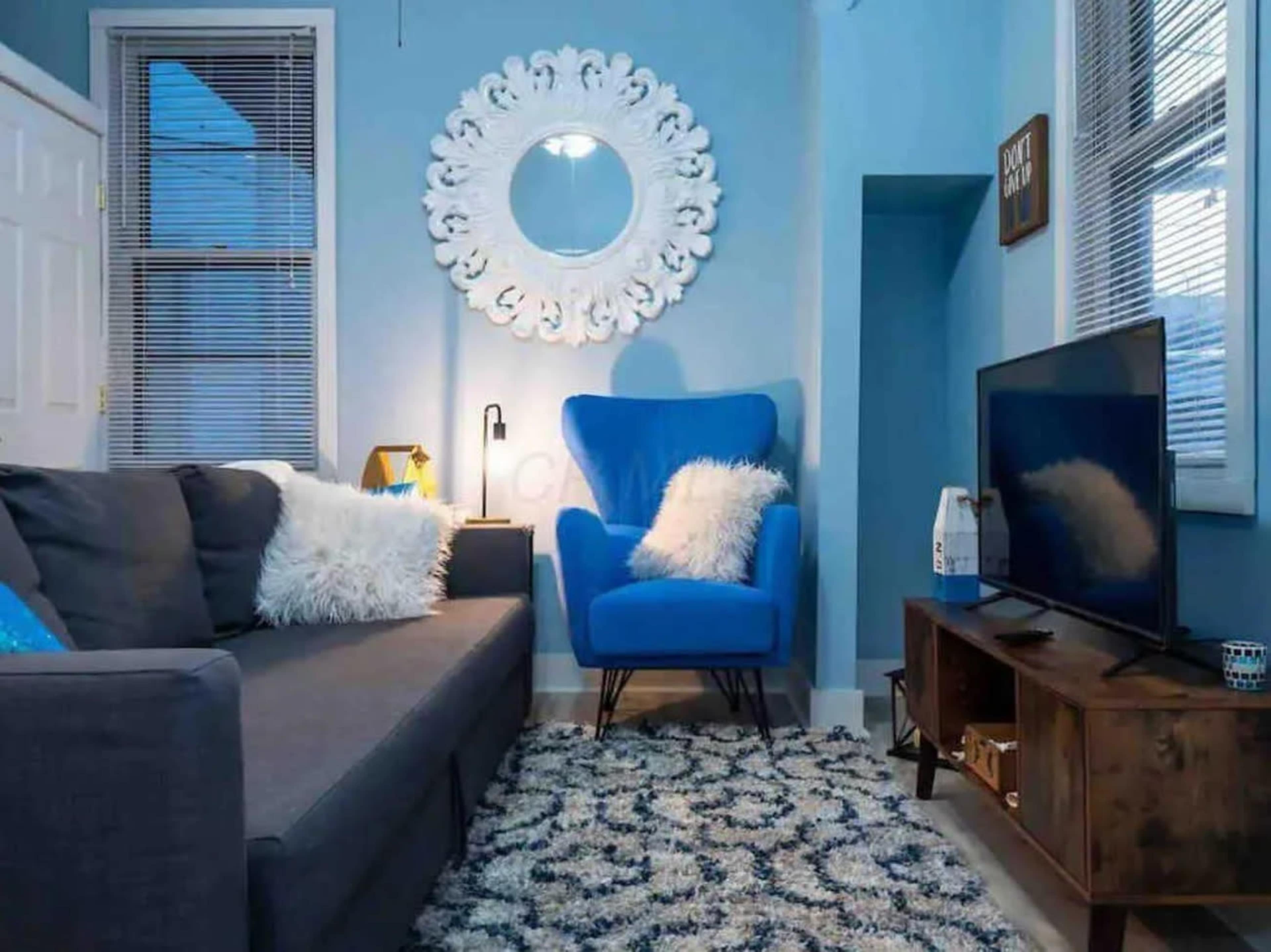 A cozy living room features a gray sectional sofa, a blue accent chair, a round mirror, and a television on a wooden stand, all set against blue walls and a patterned rug.