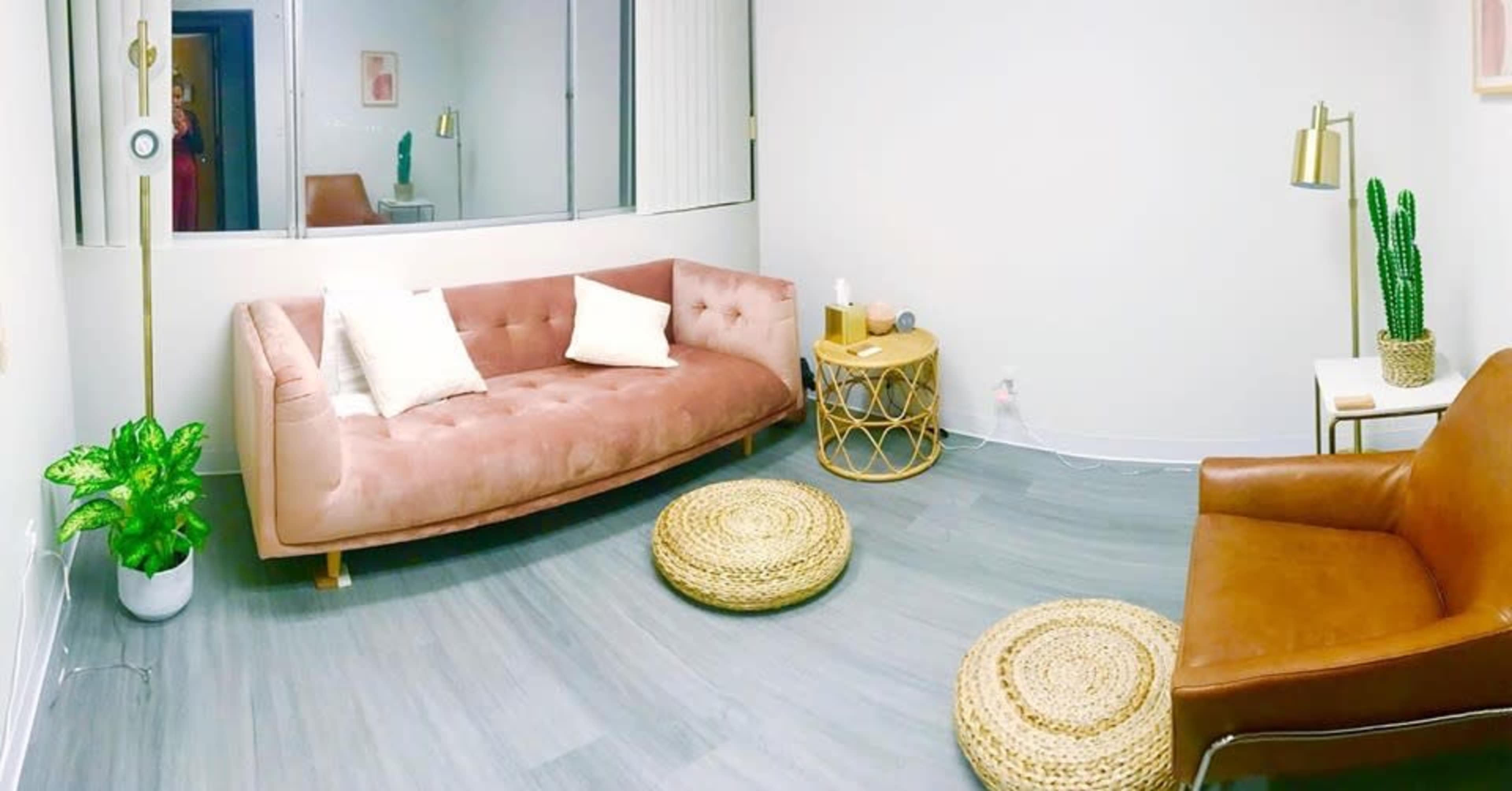 A softly lit room features a pink upholstered sofa, a brown leather chair, a small round table, and decorative plants.