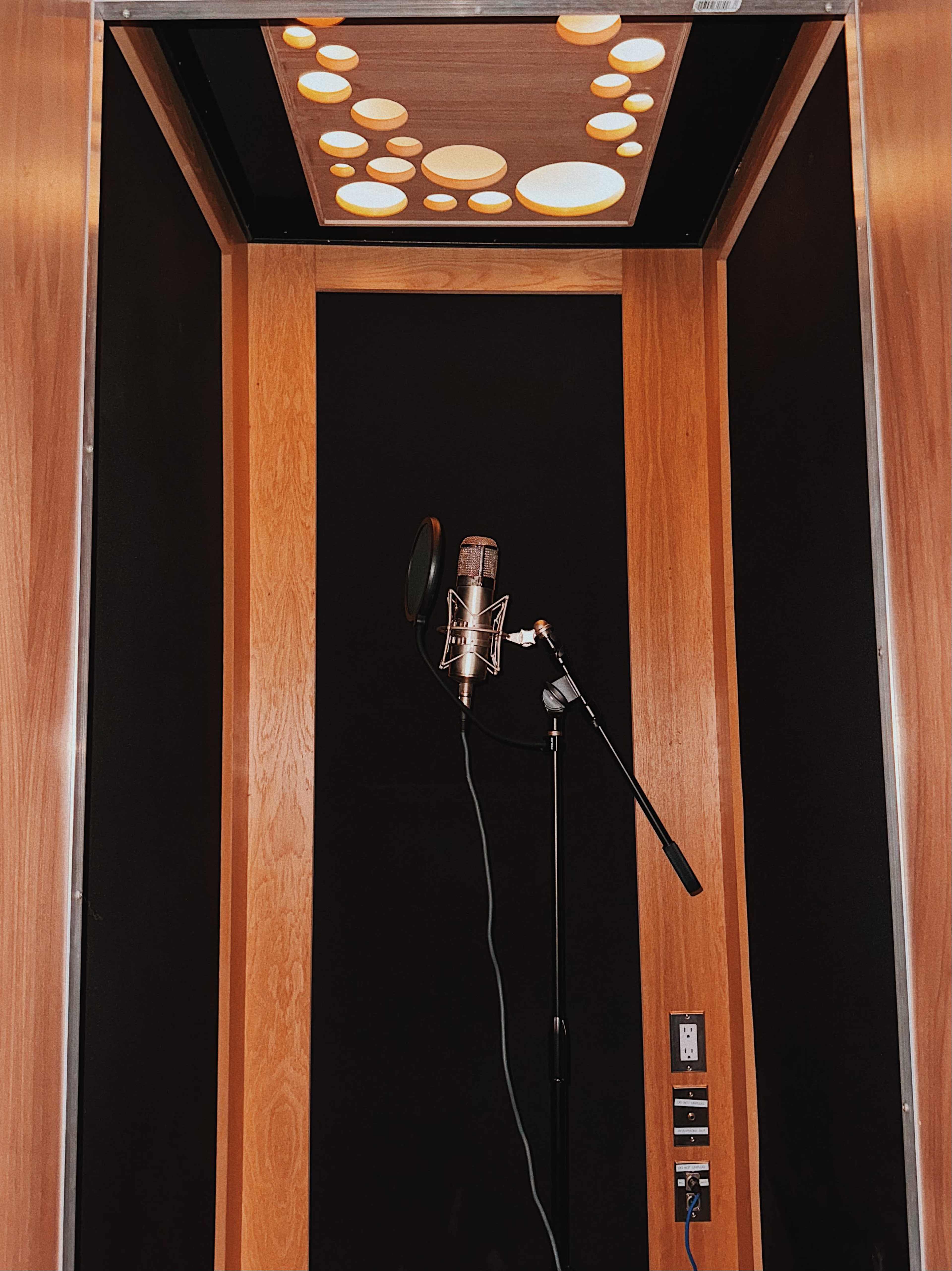 The image shows a sound booth featuring a microphone on a stand, surrounded by wooden walls and accented by circular ceiling lights.