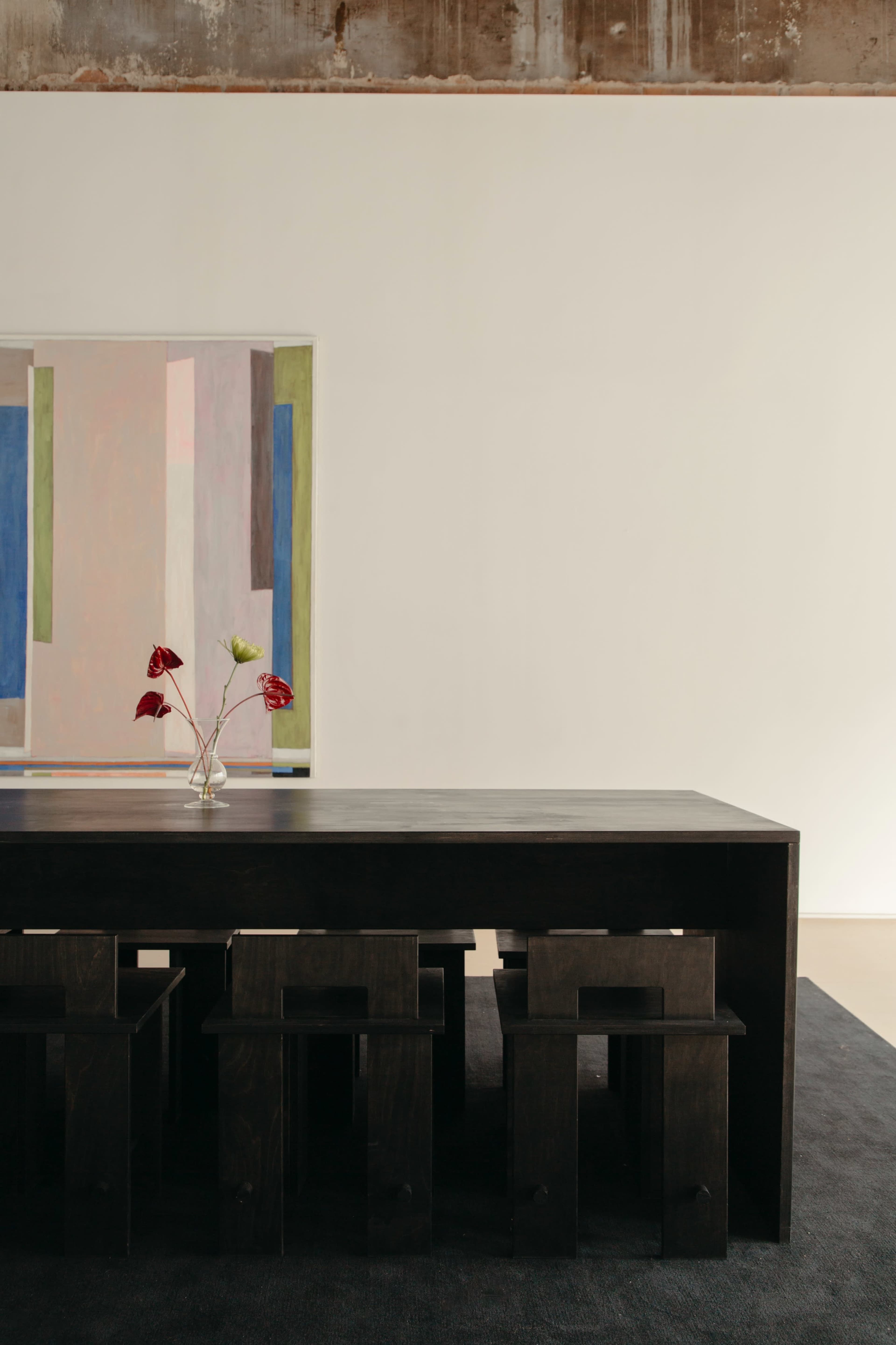 A modern dining area features a large dark wooden table and minimalist stools, complemented by a vase of flowers and an abstract painting on the adjacent wall.