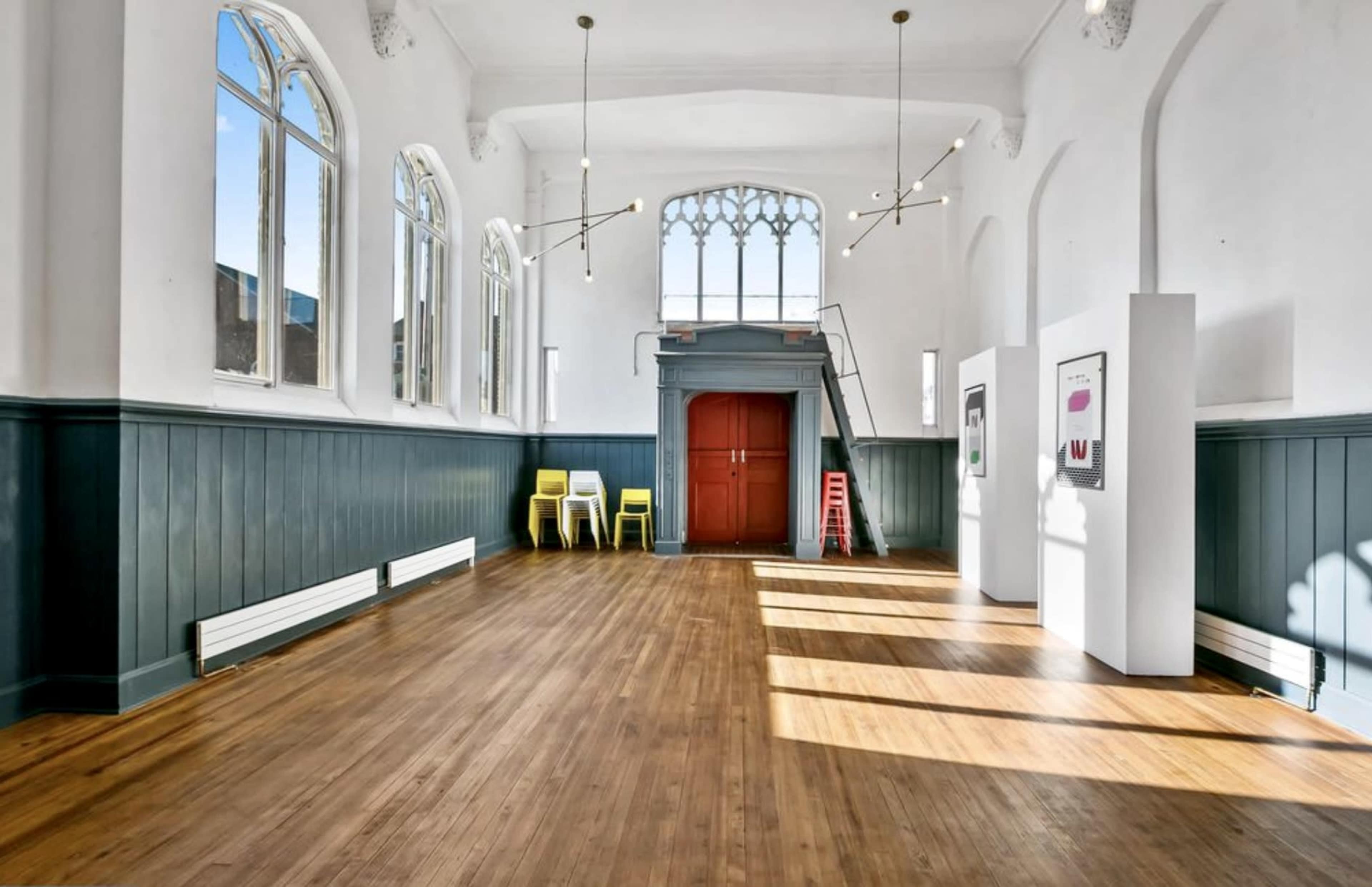 A spacious, well-lit room with wooden flooring, large windows, and minimalist furniture arranged against a backdrop of white walls and dark green paneling.