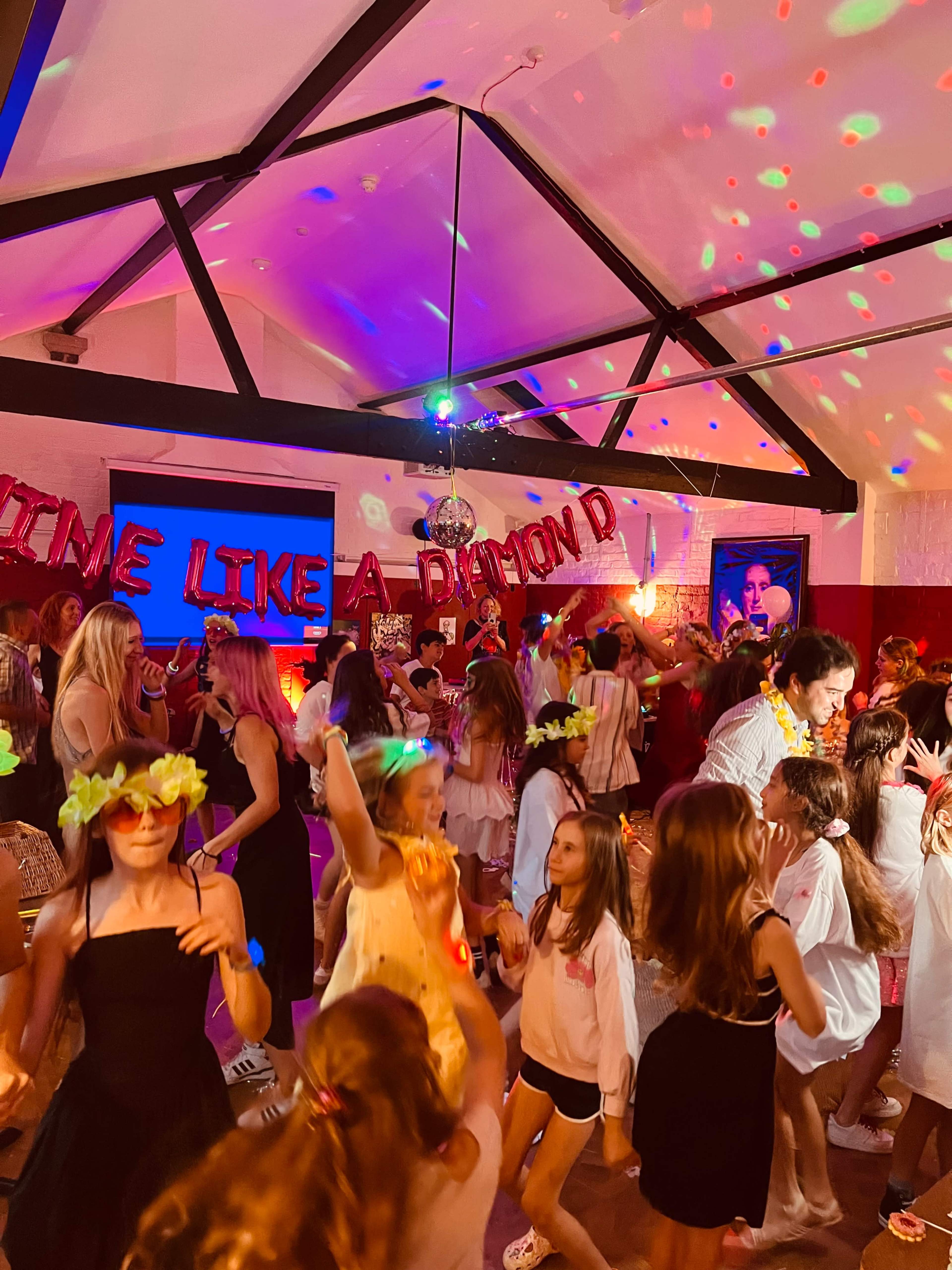 A lively group of people is dancing in a decorated room with colorful lights and a banner that reads "DANCE LIKE A DIAMOND."