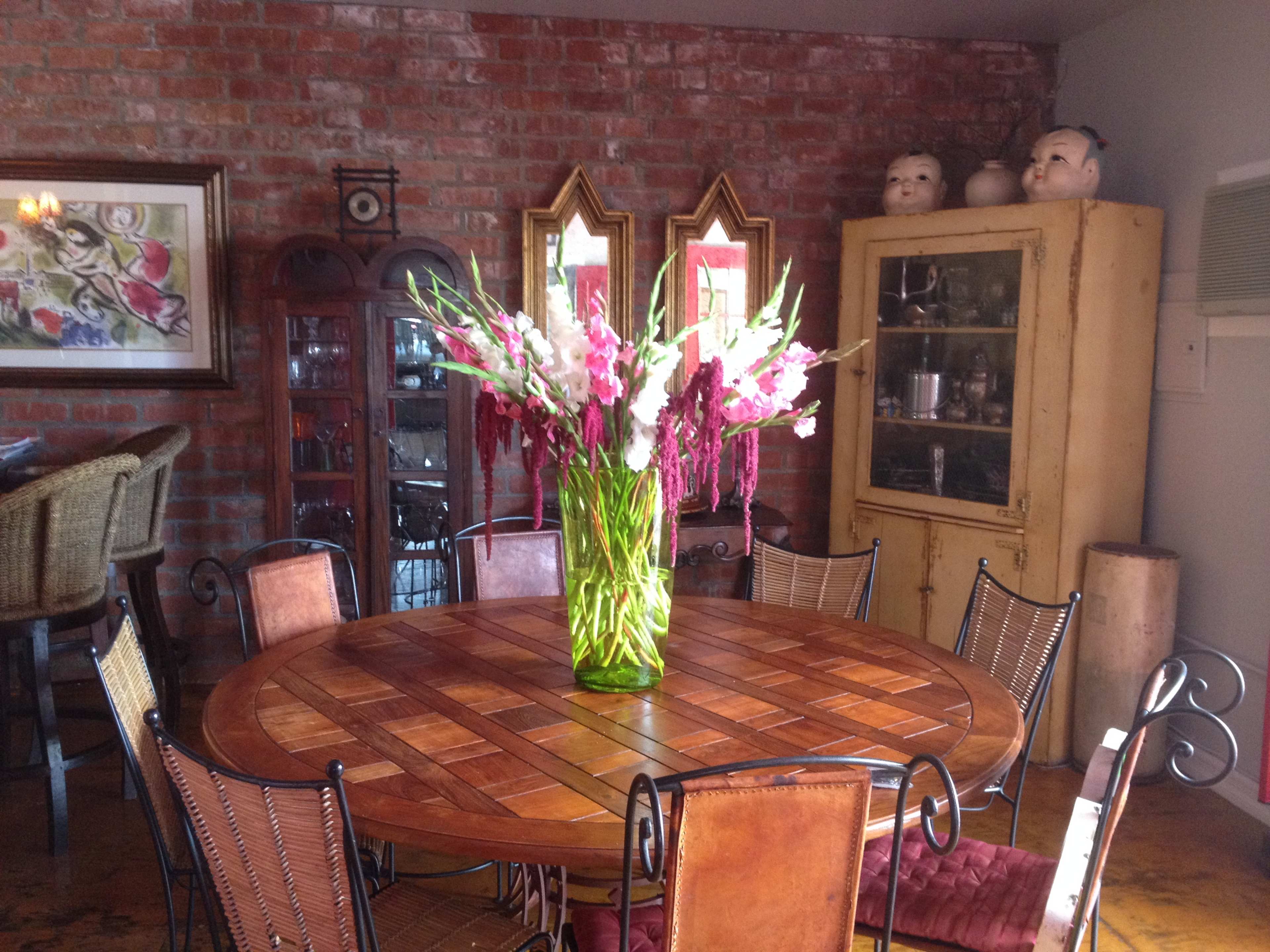 A round wooden dining table is surrounded by wrought iron chairs, topped with a large vase of flowers in a brick-walled room featuring a cabinet and decorative mirrors.