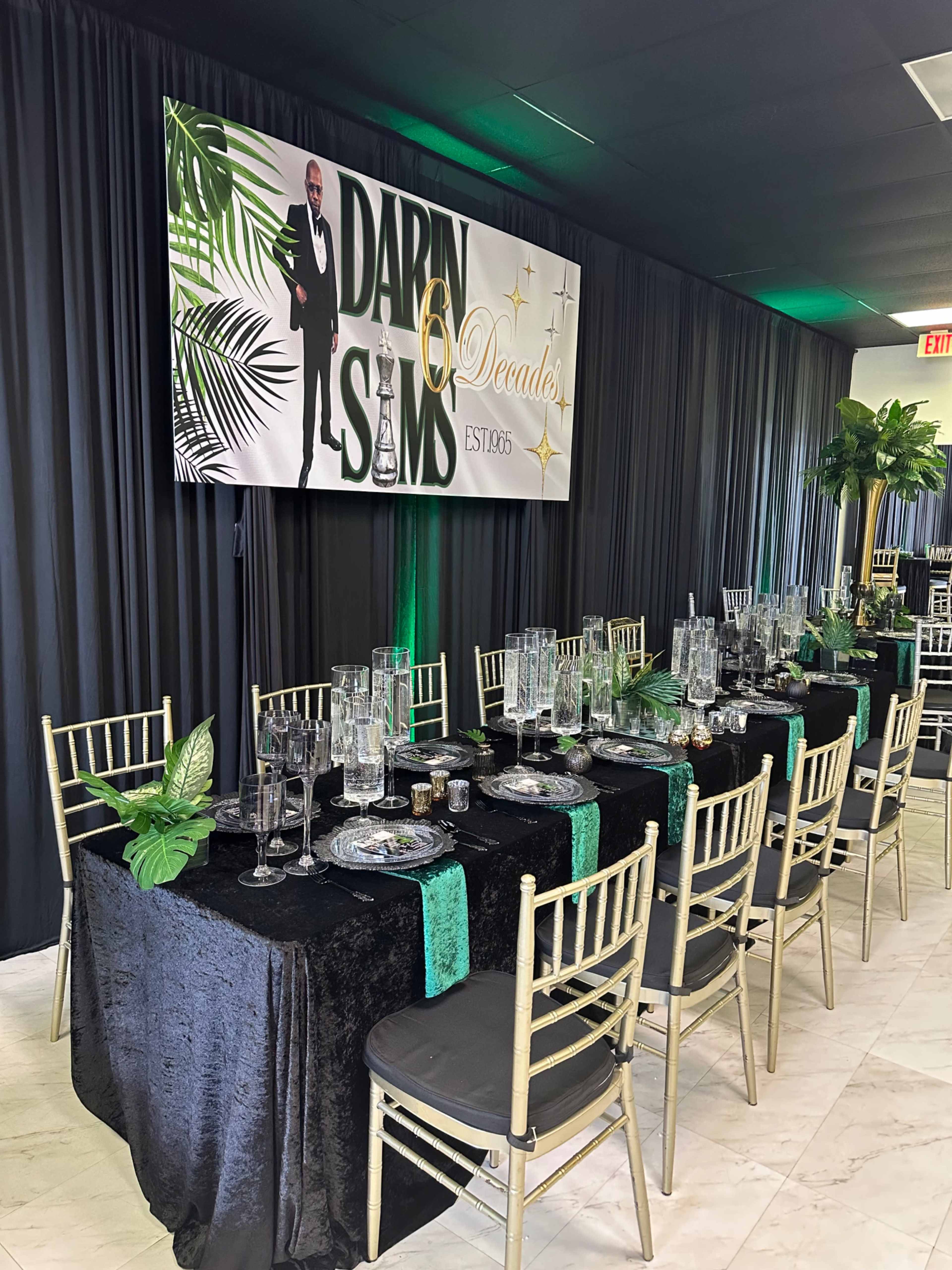 The image shows a decorated banquet table set with elegant tableware, surrounded by black curtains and a large sign celebrating "Darin's 60th Decades."