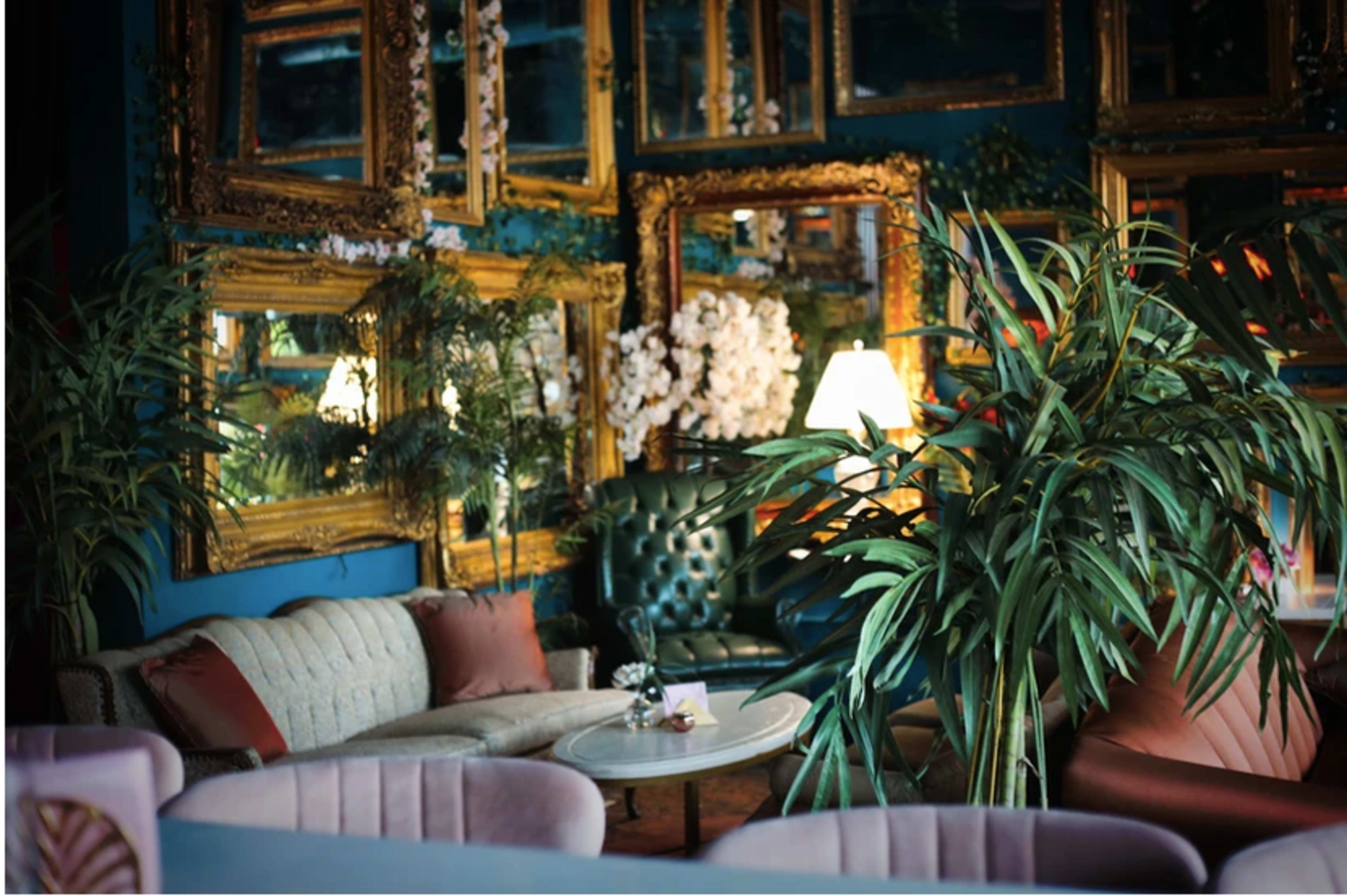 The scene features a stylish interior with multiple ornate mirrors, green plants, and vintage furniture arranged around a small table.