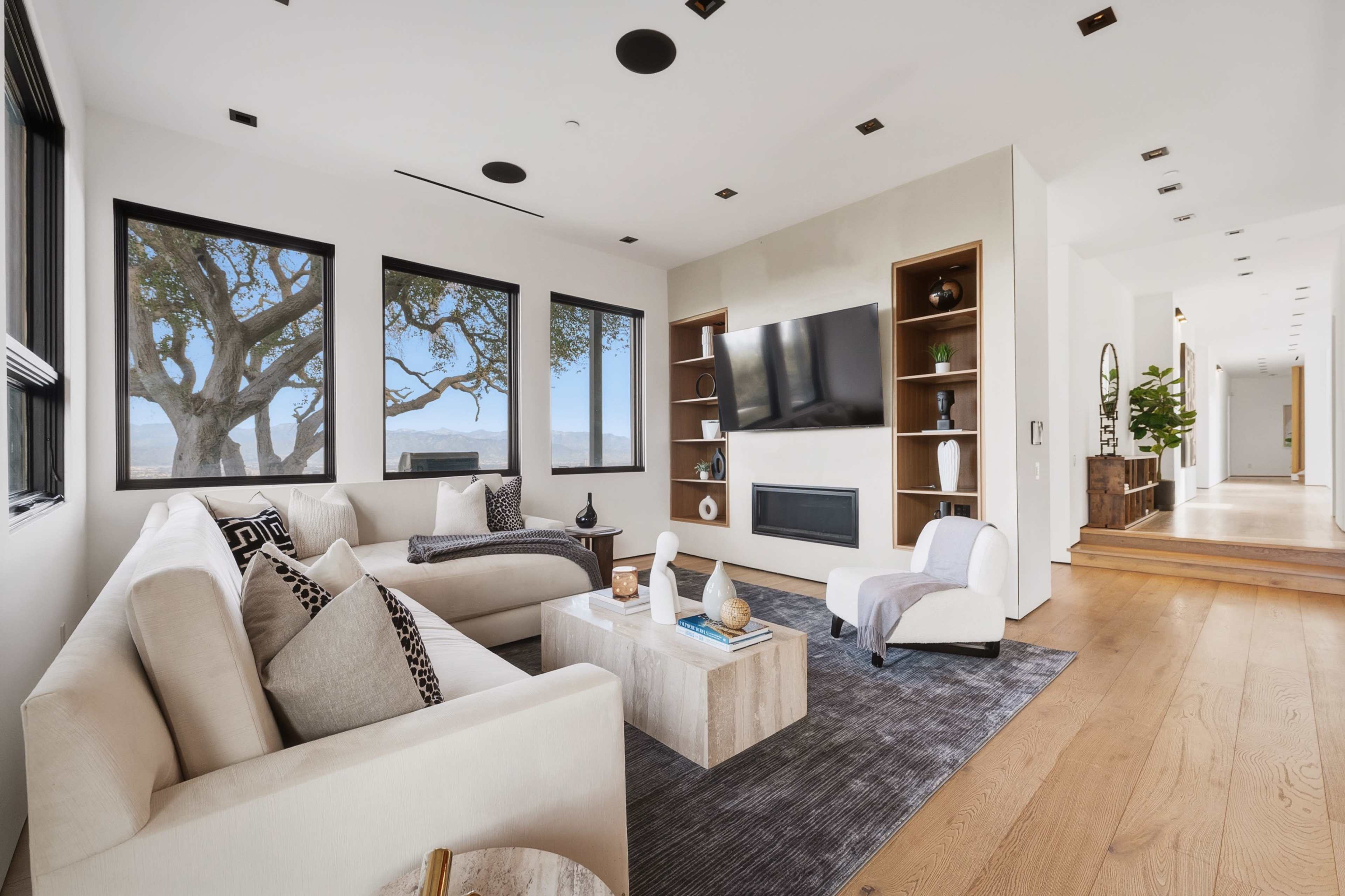 A modern living room features a sectional sofa, a coffee table, and a wall-mounted television, with large windows providing views of a tree outside.