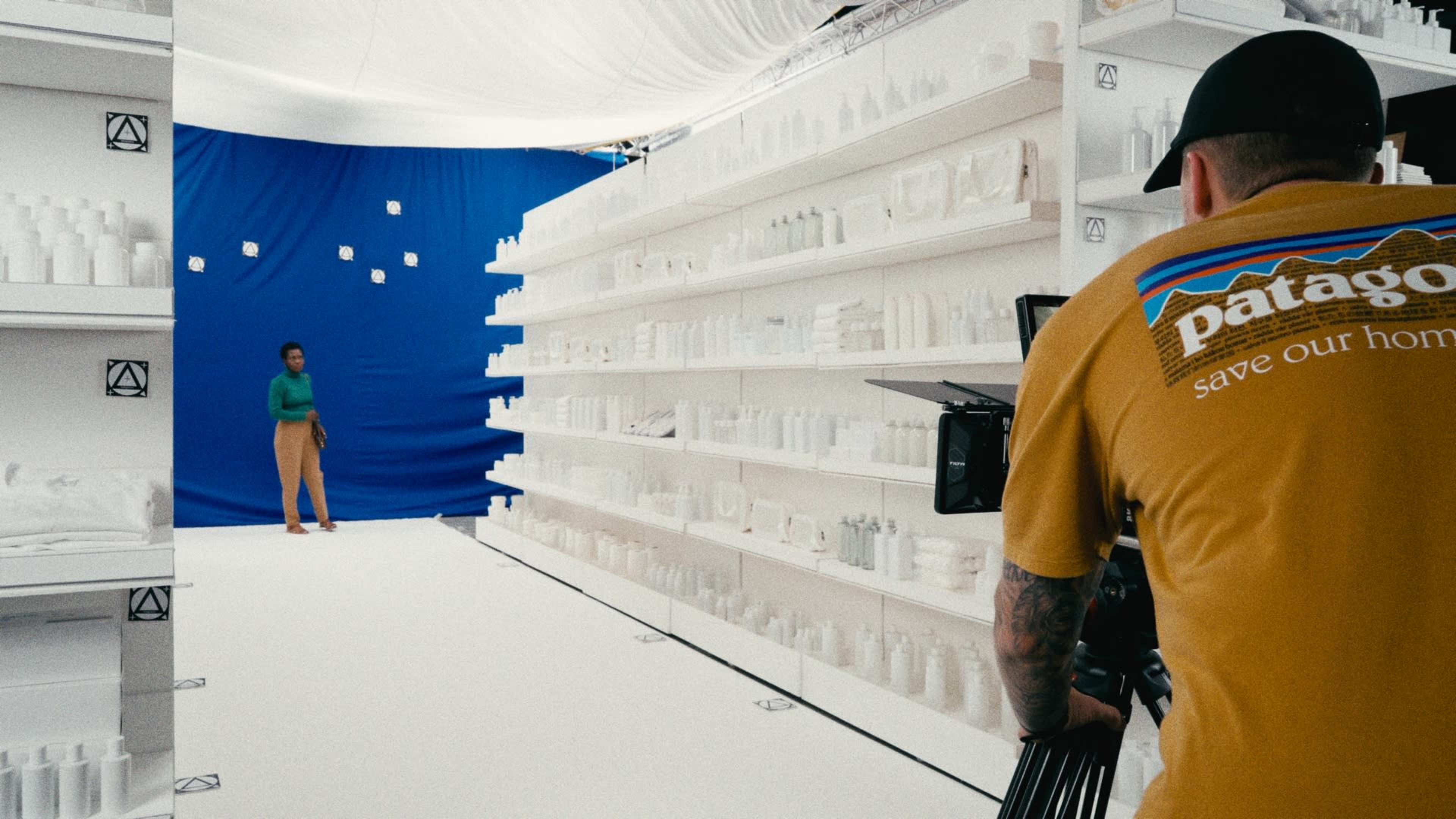 A person stands on a white set with shelves displaying various bottles, while a camera is positioned behind an operator wearing a yellow Patagonia shirt, against a blue backdrop.