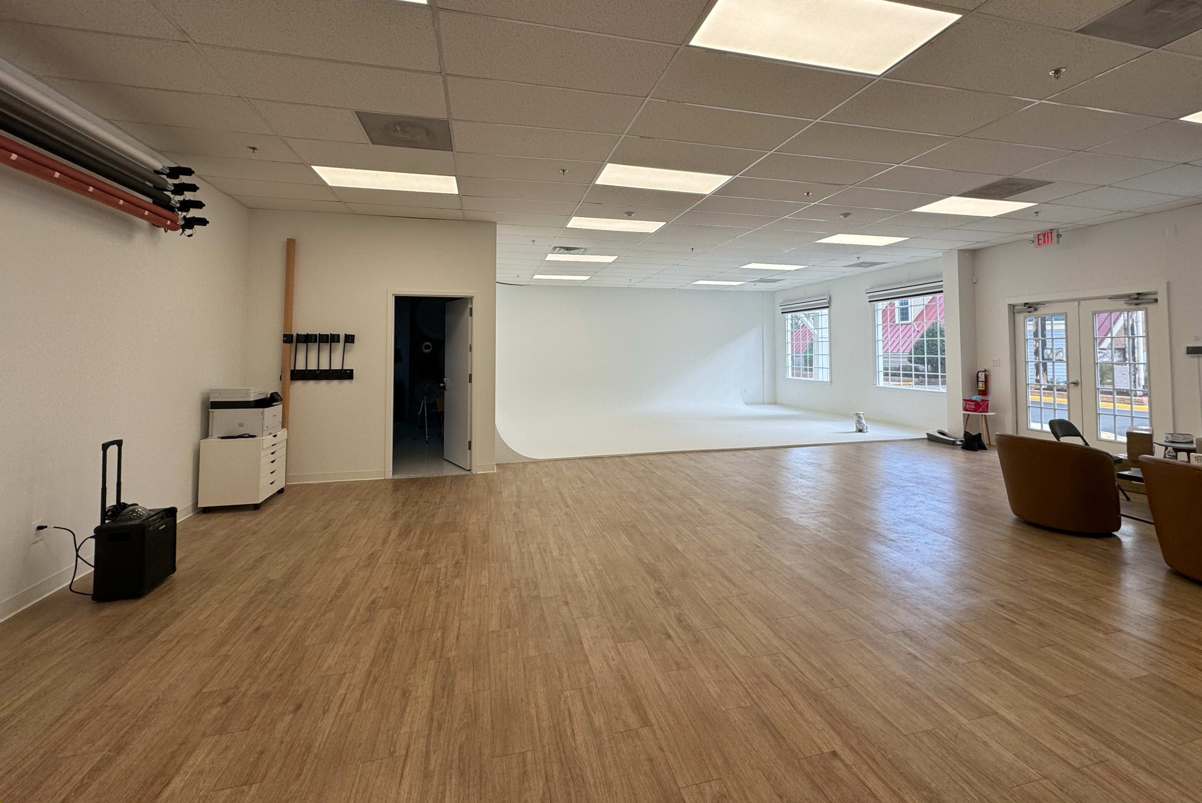 A spacious, well-lit studio with a seamless white backdrop, wooden flooring, and minimal furniture on one side.