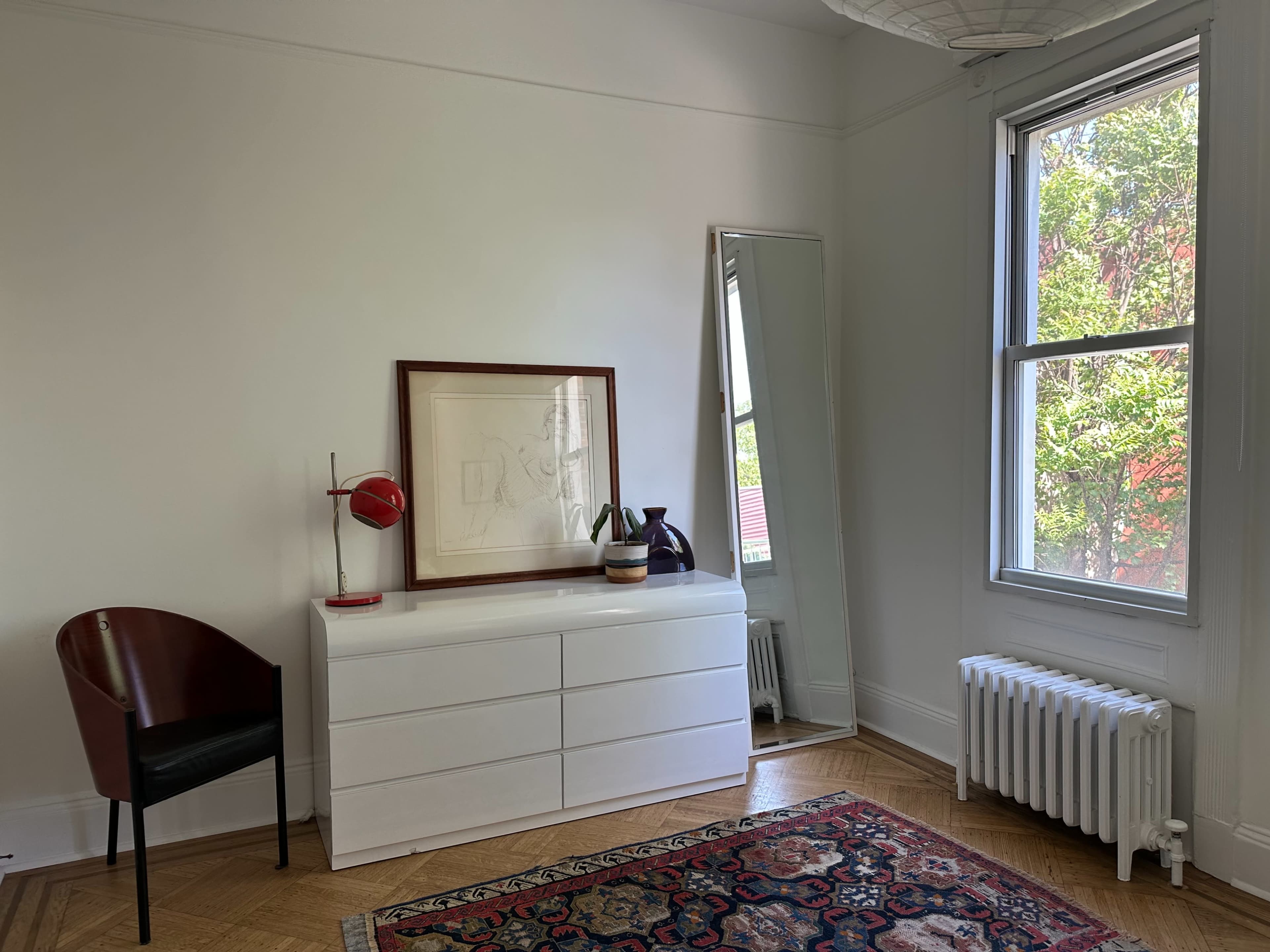 The image shows a minimalistic bedroom with a white dresser, a large mirror, and a chair next to a window, displaying a radiator and some greenery outside.
