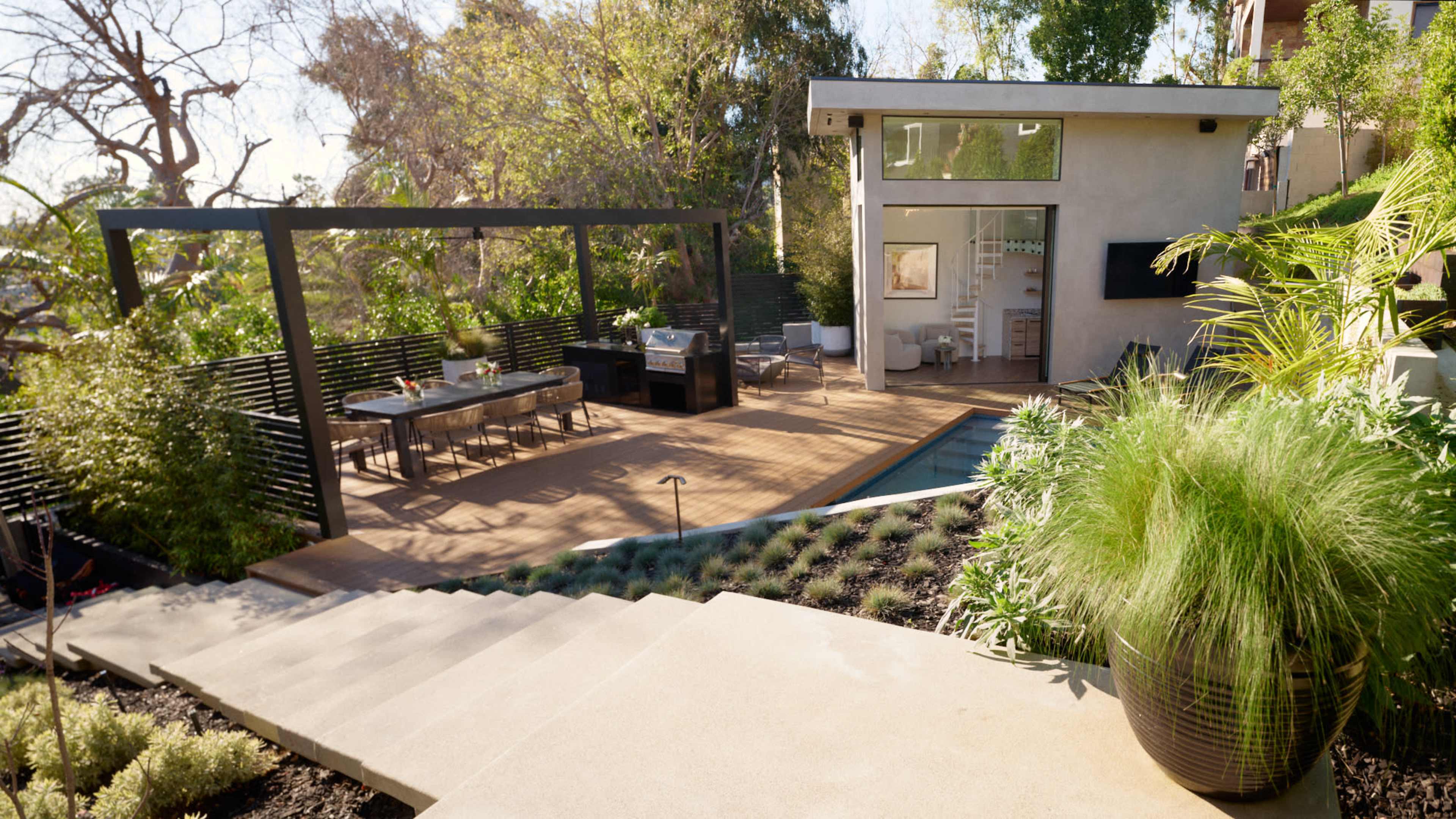 A modern backyard featuring a wooden deck with outdoor furniture, a barbecue area, and a swimming pool, surrounded by landscaped greenery.