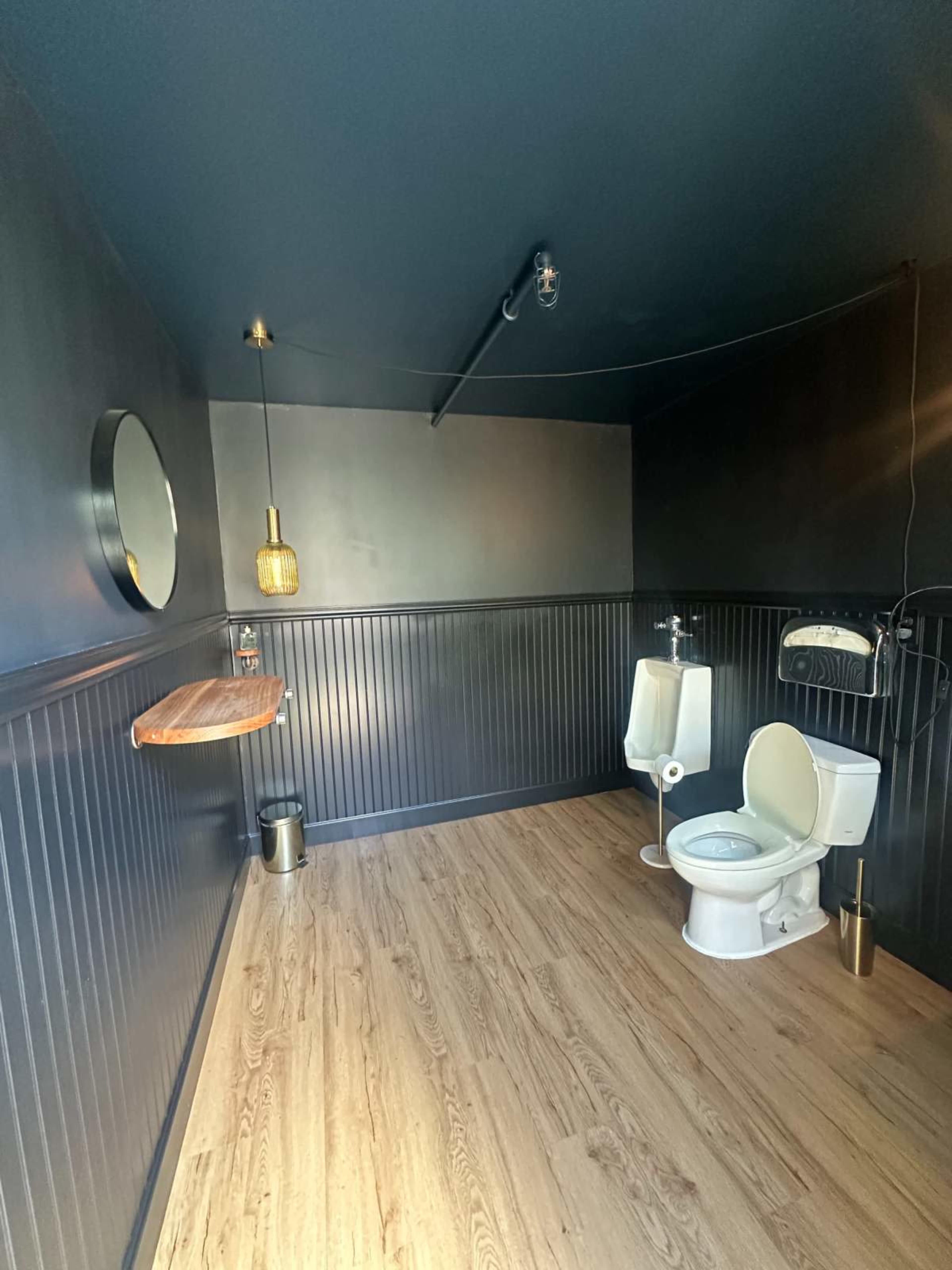 A small restroom featuring a toilet, two urinals, a round mirror, and a wooden shelf against a dark wall.