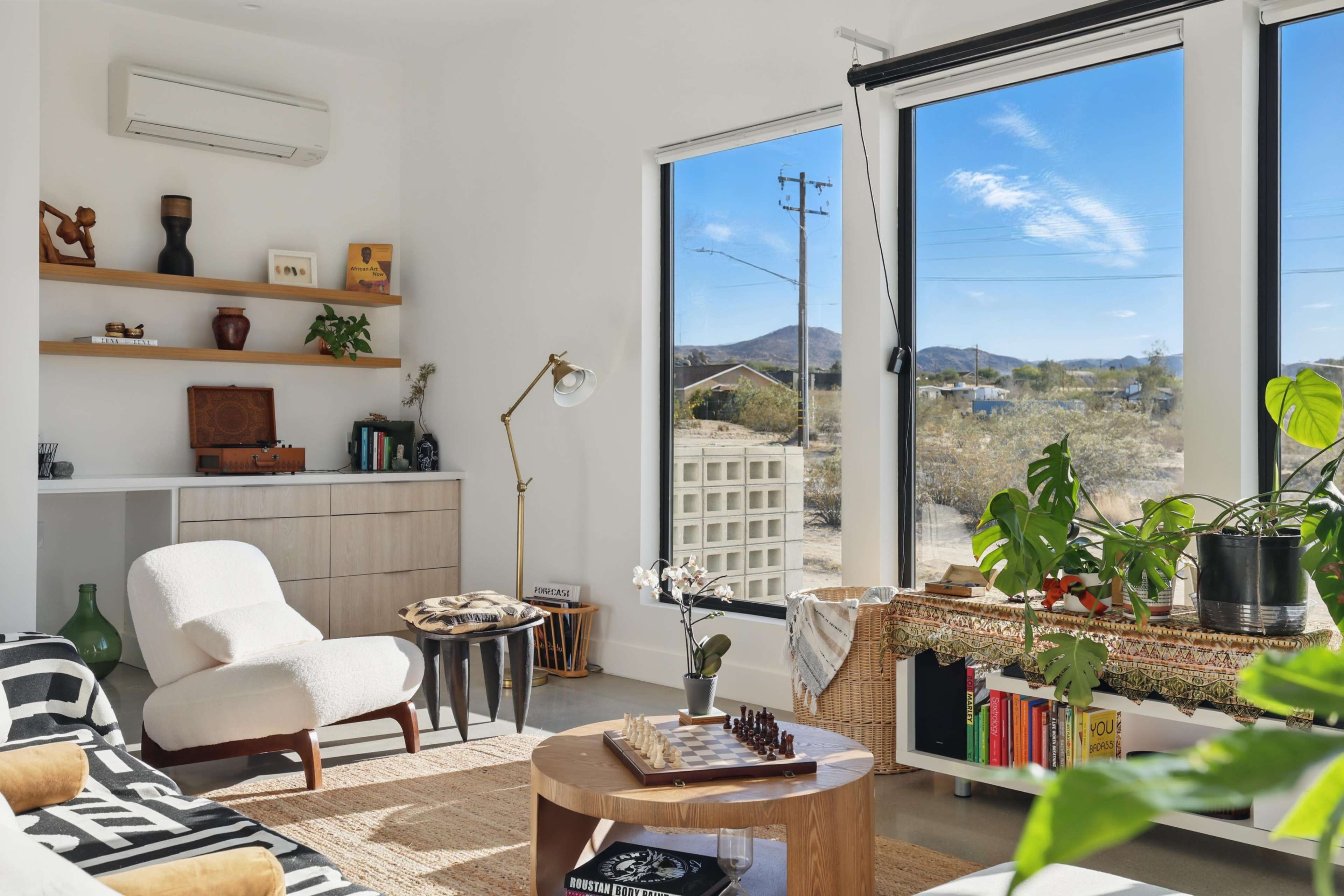 A modern, sunlit living room features large windows, a plush armchair, a wooden coffee table, and a variety of houseplants.