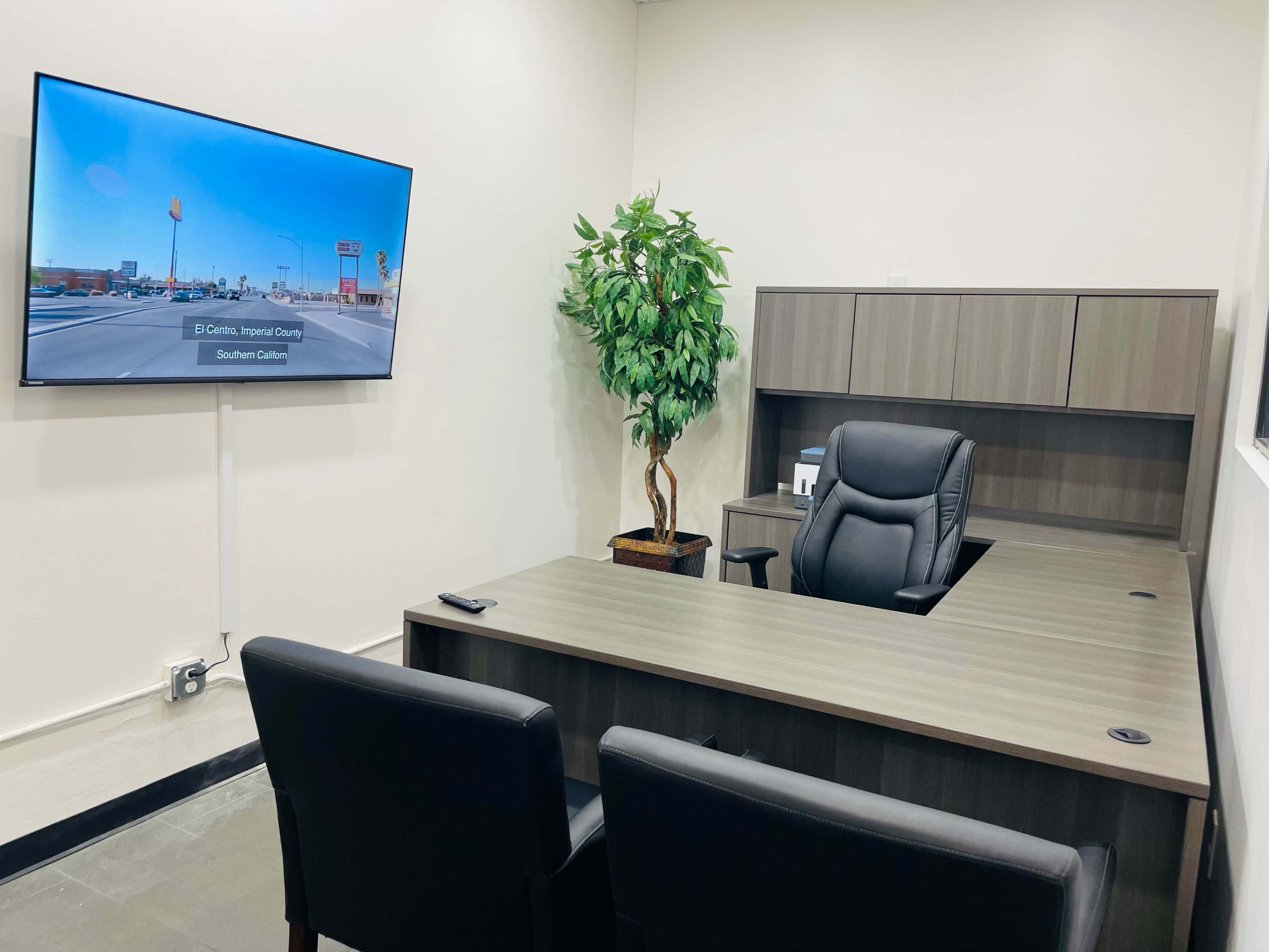The image shows a modern office with a large desk, an office chair, two black chairs, a potted plant, and a mounted television displaying a road scene.