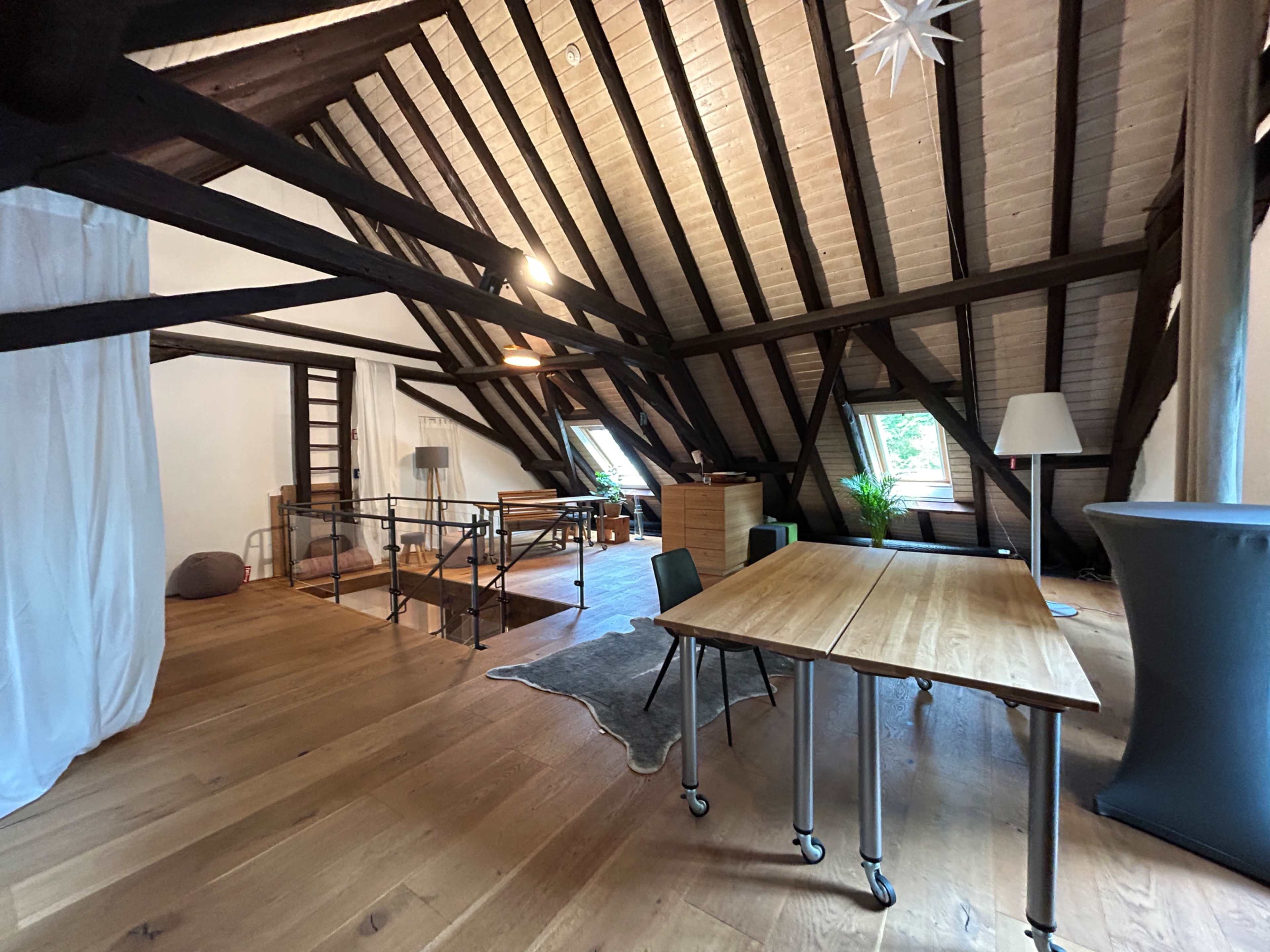 The image shows an attic interior featuring wooden beams, a wooden floor, a workspace with tables, and a small seating area.