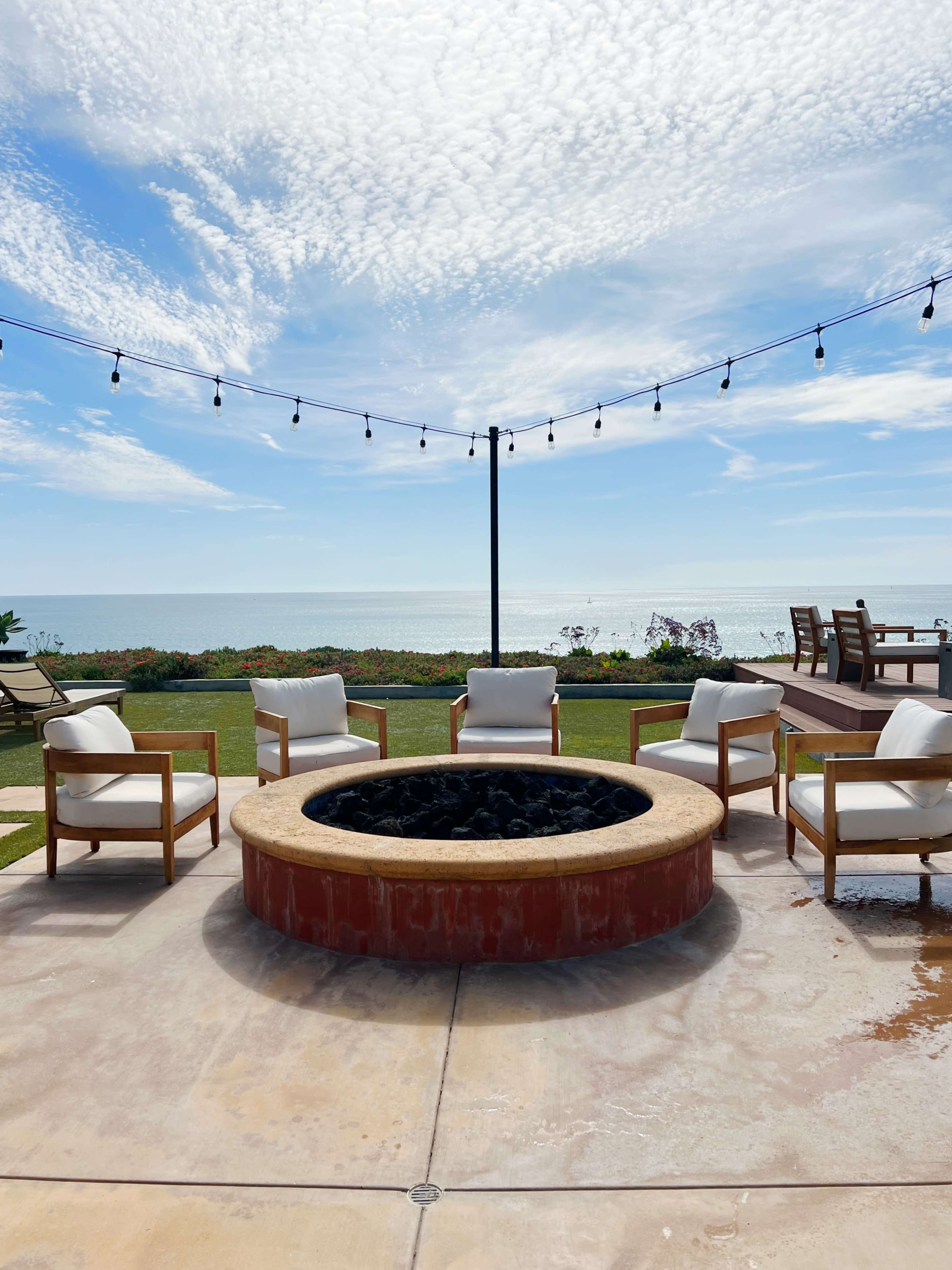 A circular fire pit surrounded by four white chairs is positioned near a coastline with a view of the ocean and a cloudy sky overhead.