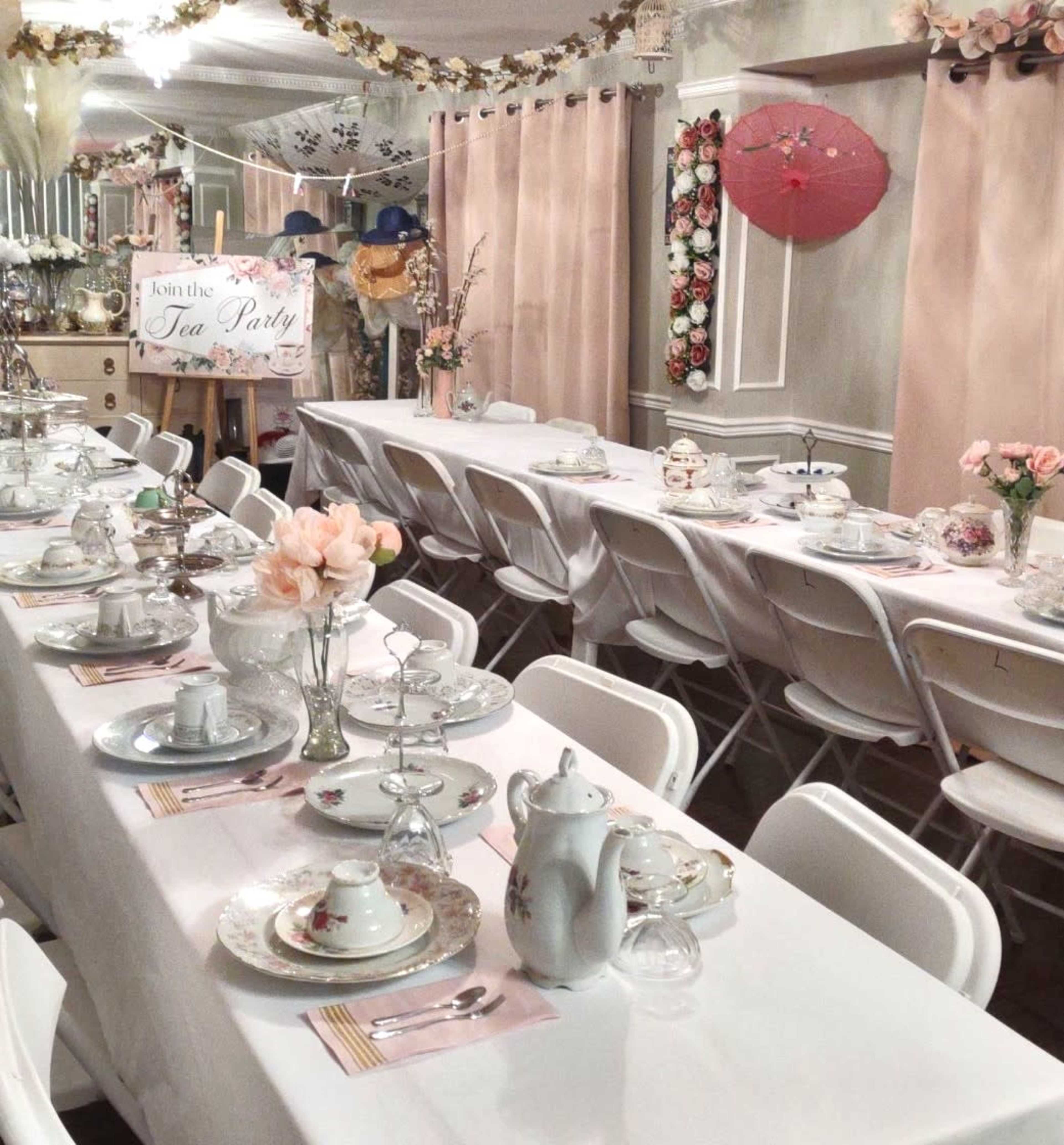 A long table is elegantly set for a tea party, adorned with fine china, floral arrangements, and decorative elements in a softly lit room.
