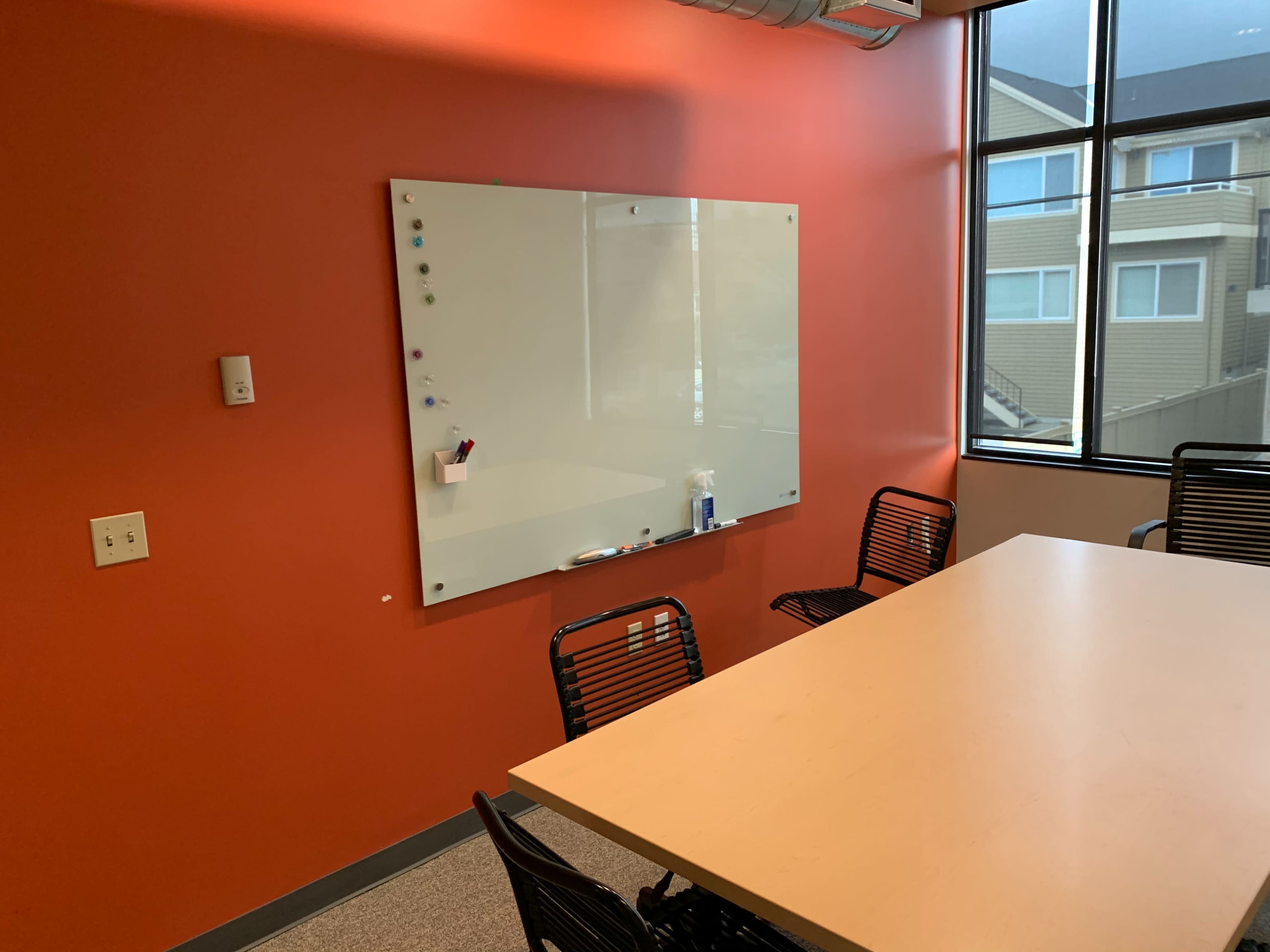 A conference room with a large whiteboard on an orange wall and a light-colored table surrounded by black chairs.