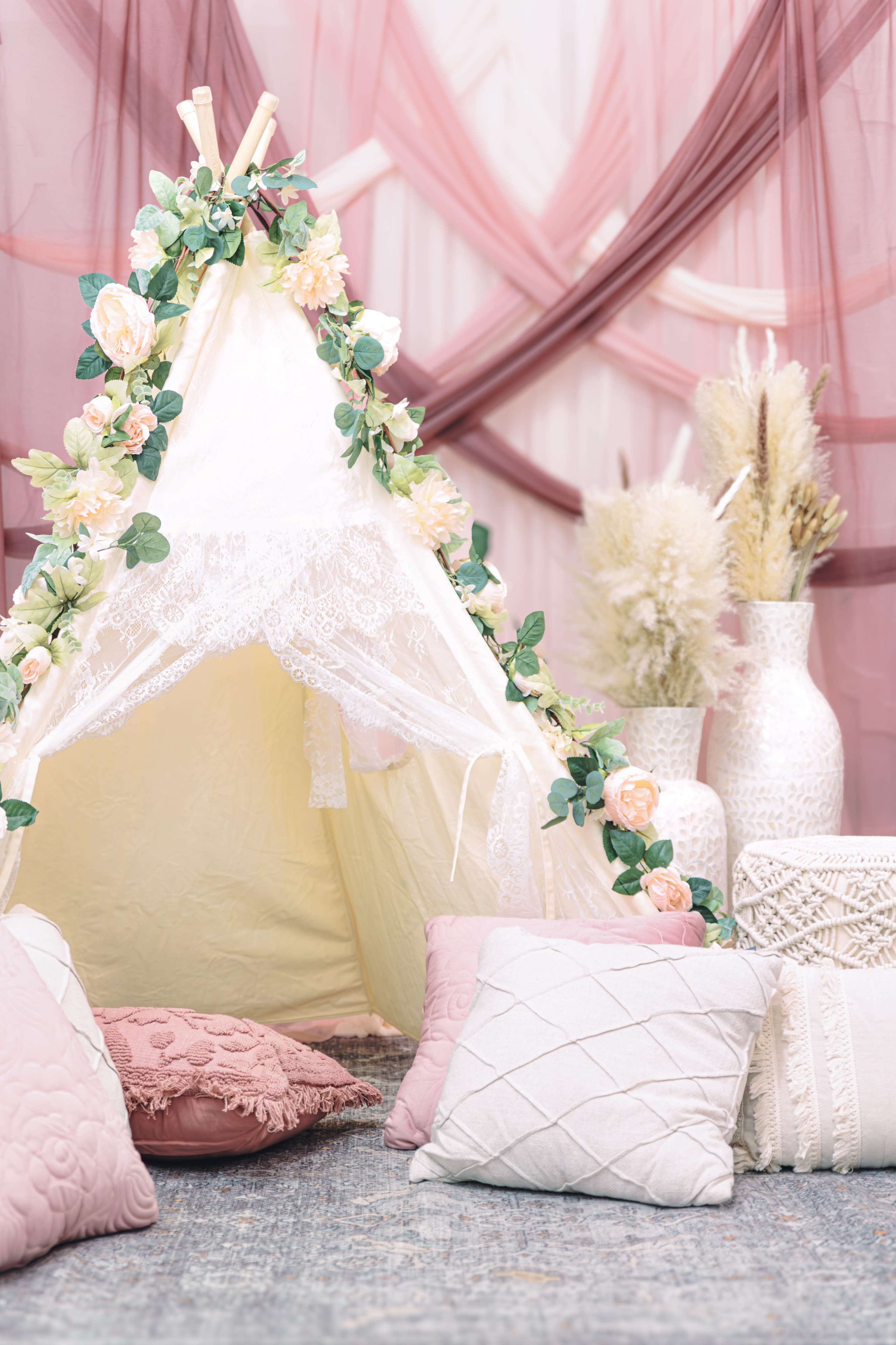 The image shows a decorative tent made of fabric, adorned with flowers, surrounded by soft pillows and vases in a cozy indoor setting.