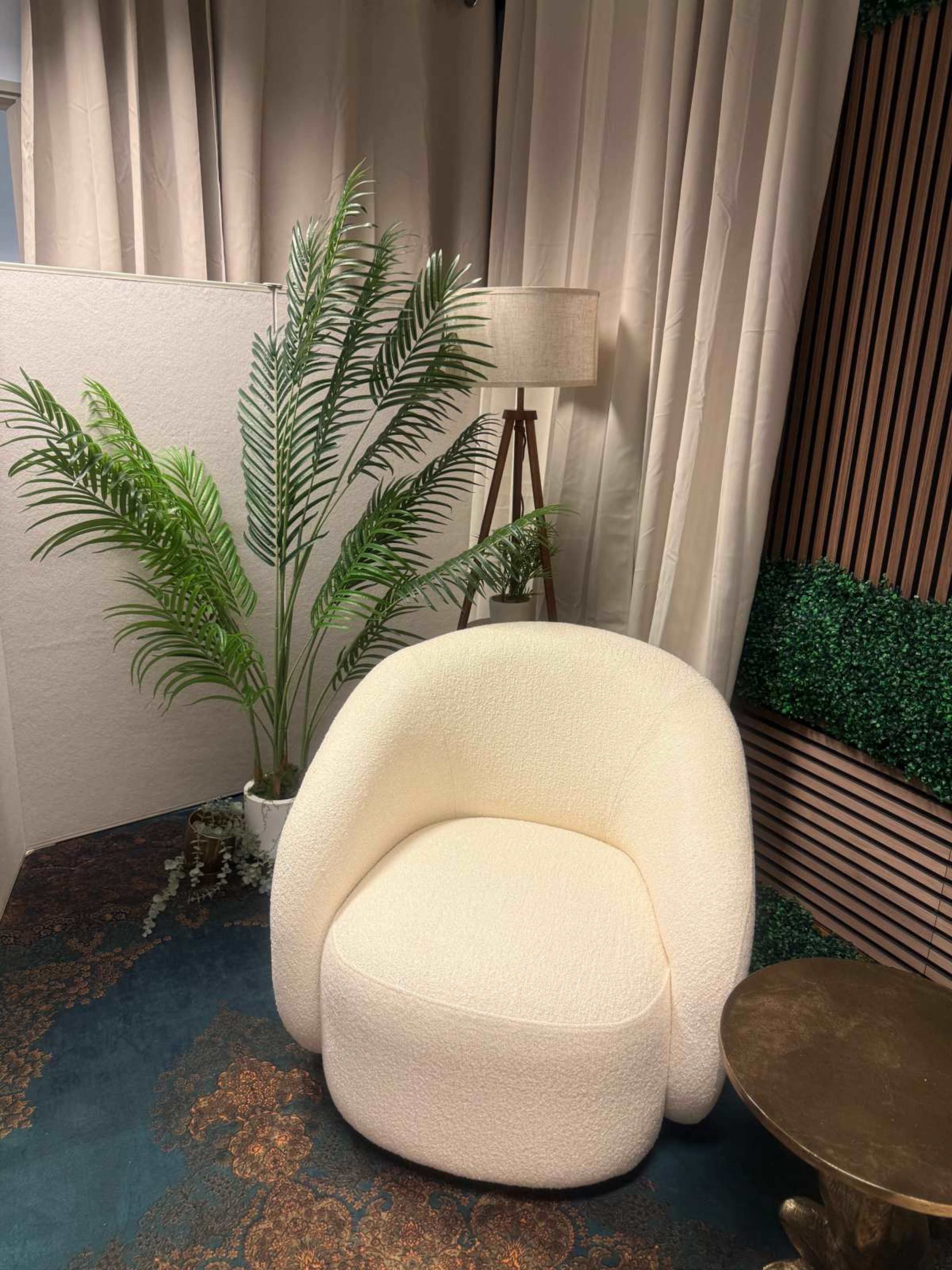The image shows a rounded, cream-colored chair beside a tall plant in a pot, with a lamp and textured wall in the background.
