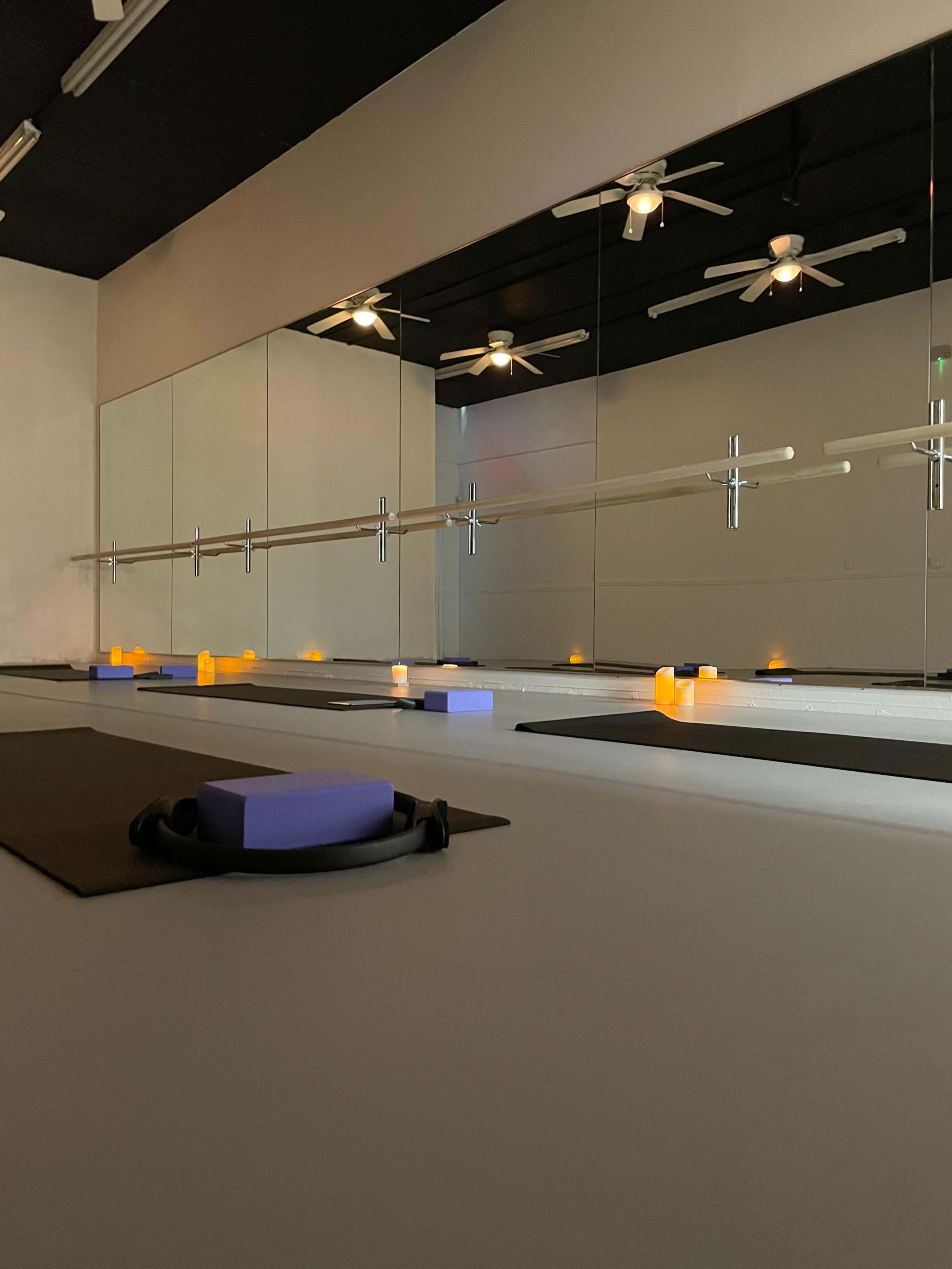 The image shows a spacious, minimalistic yoga studio with mirrored walls, exercise mats laid out, and soft lighting from candles.