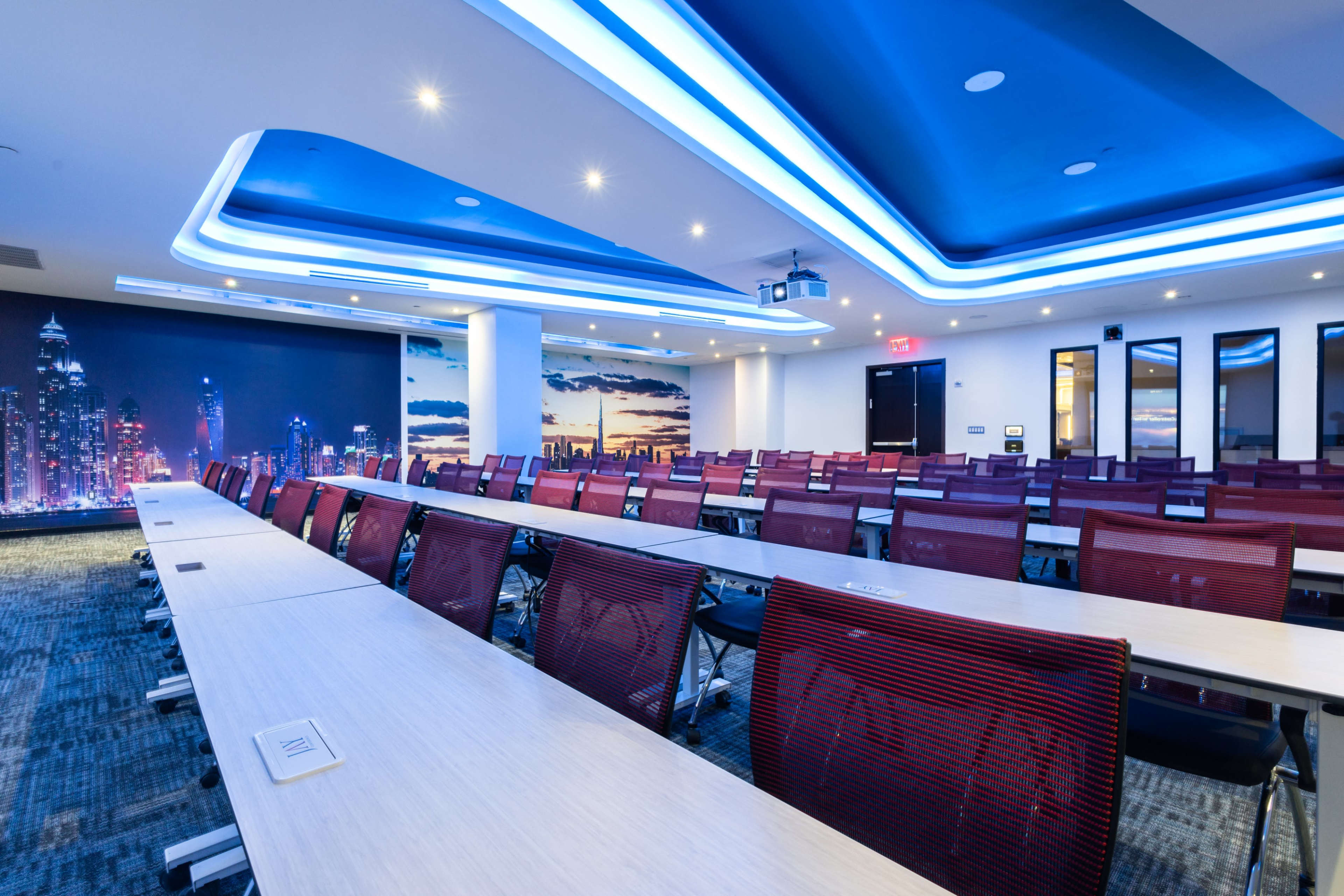 A modern conference room with rows of tables and chairs arranged neatly, illuminated by blue lighting and featuring a skyline mural on one wall.