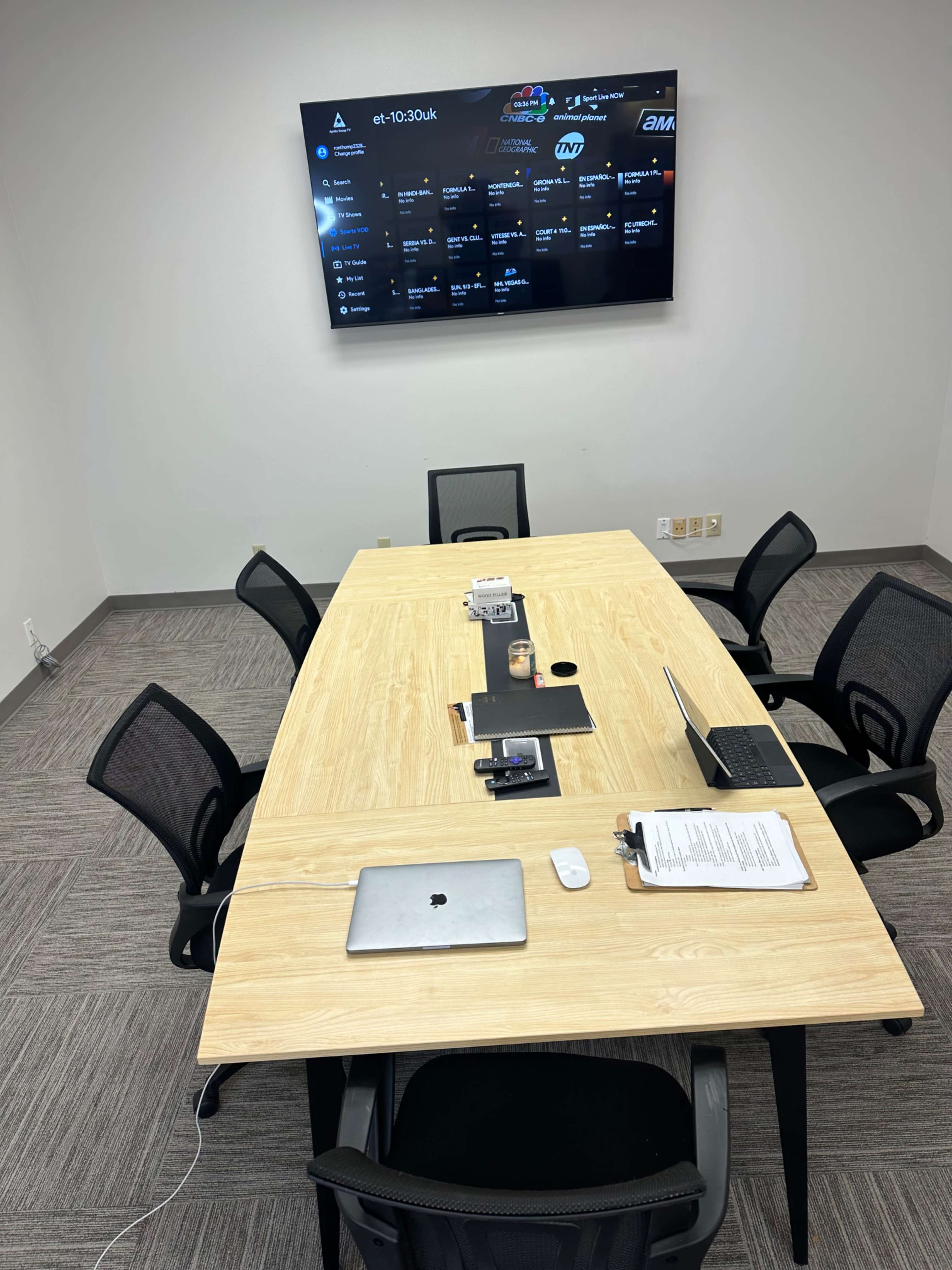 A conference room with a wooden table surrounded by six black chairs, featuring a mounted screen displaying a schedule and a laptop on the table.