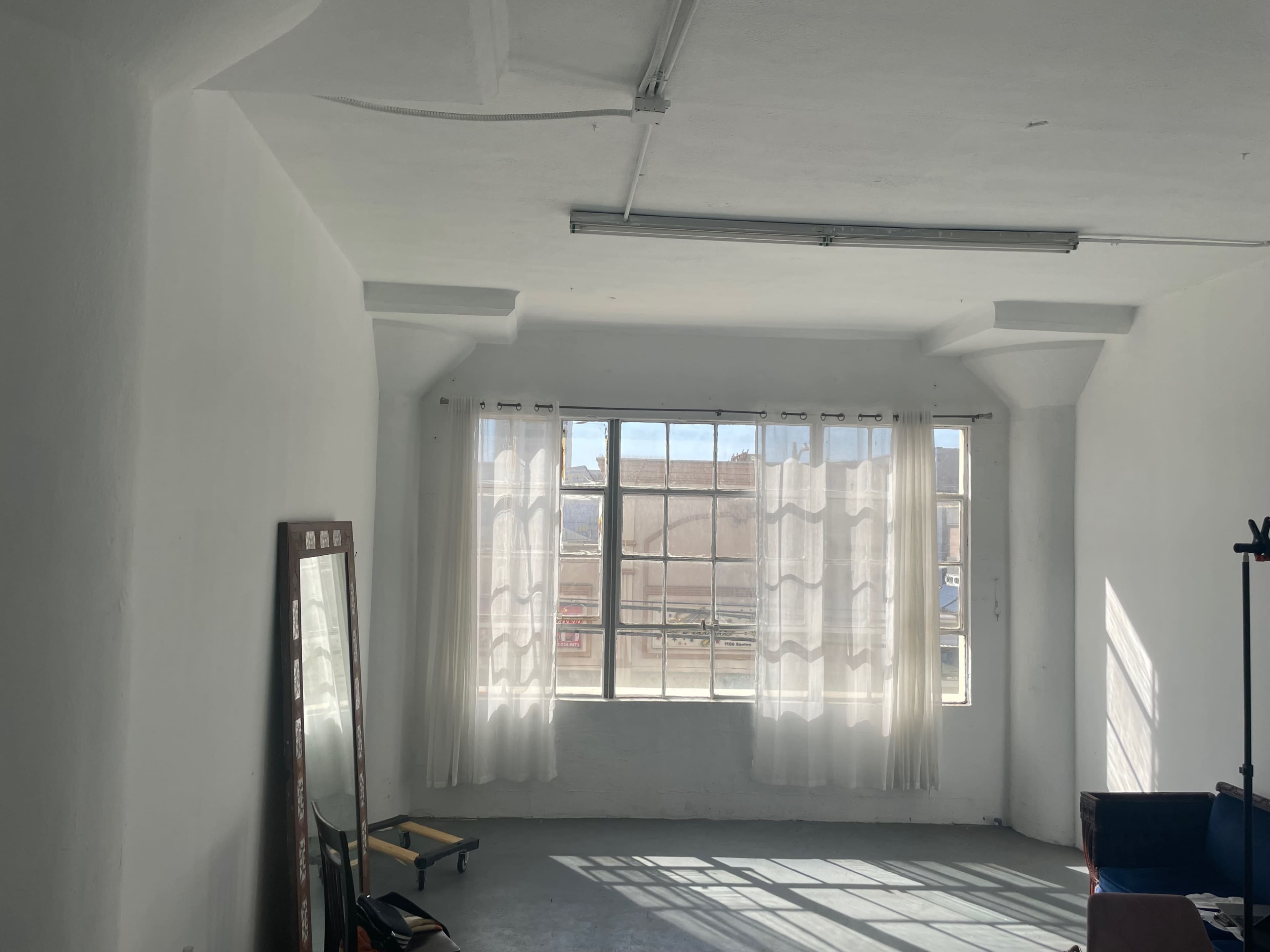 A bright, empty room features large windows with sheer curtains and a mirror on the wall.