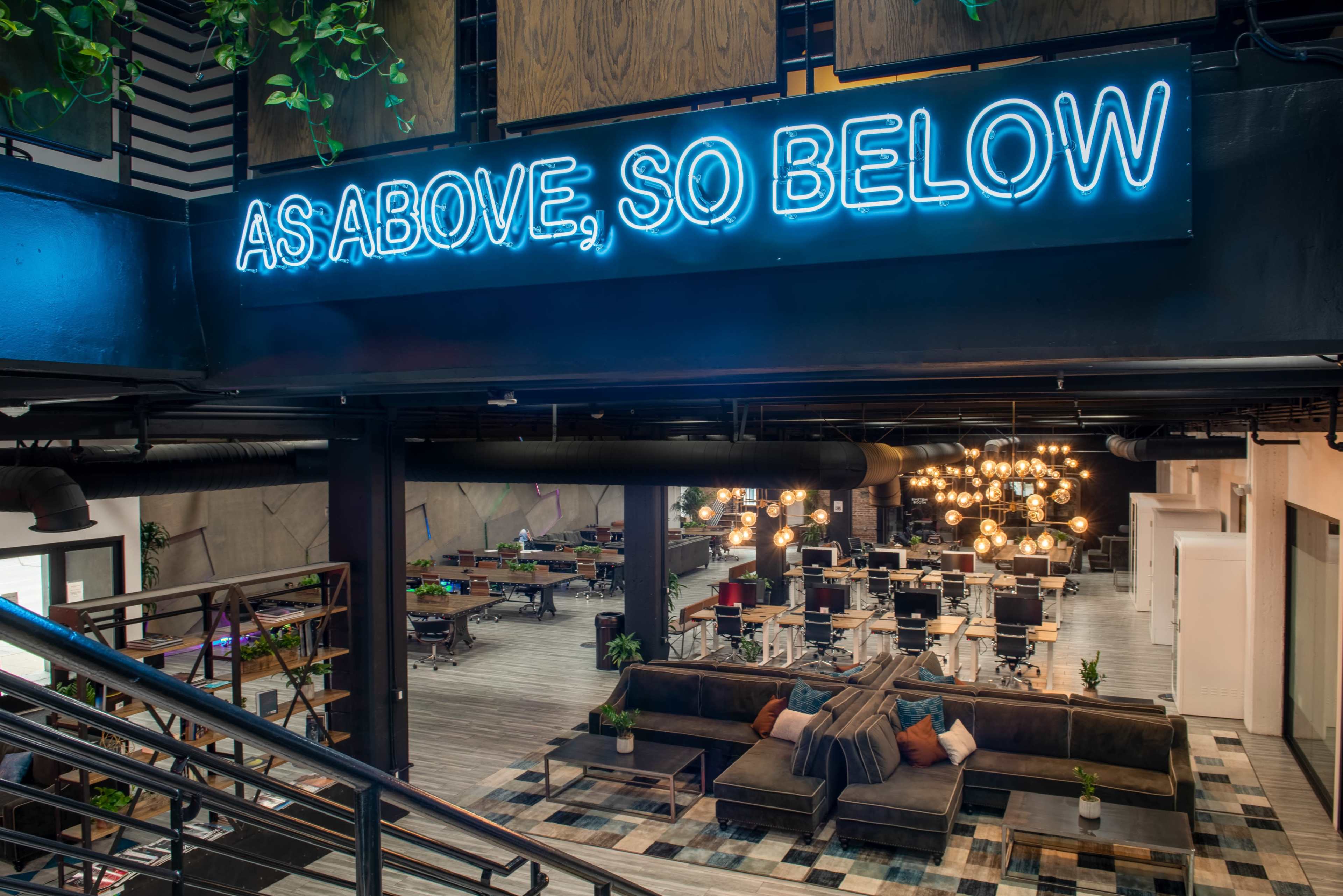 A modern indoor space with a neon sign that reads "AS ABOVE, SO BELOW," overlooking a communal area furnished with couches and tables.