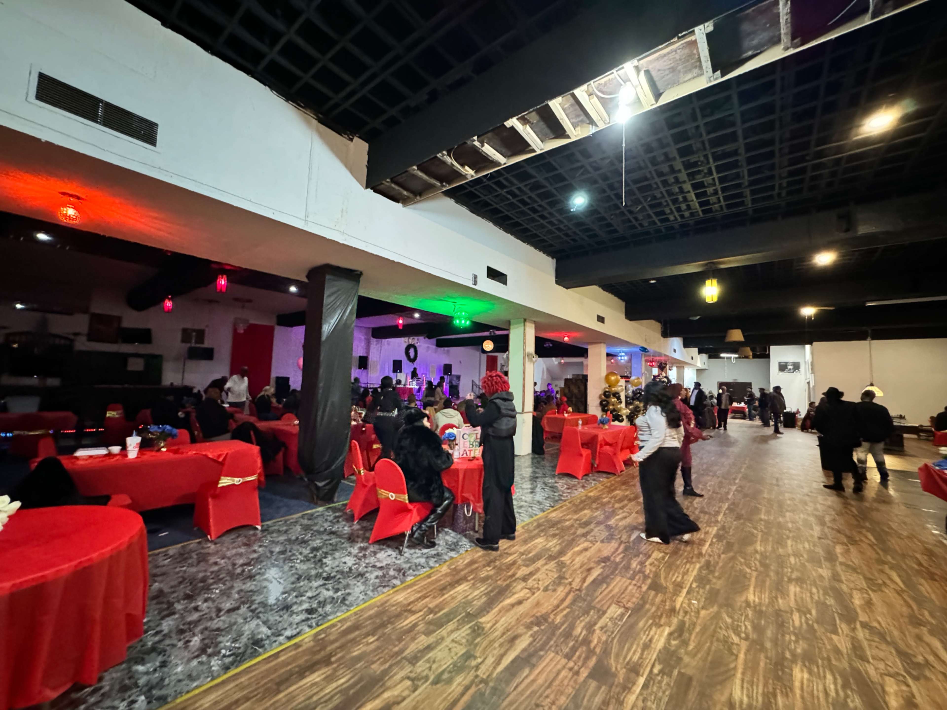 A spacious event hall is filled with tables covered in red tablecloths and attendees in formal attire, while a stage at the back is set up for a performance.