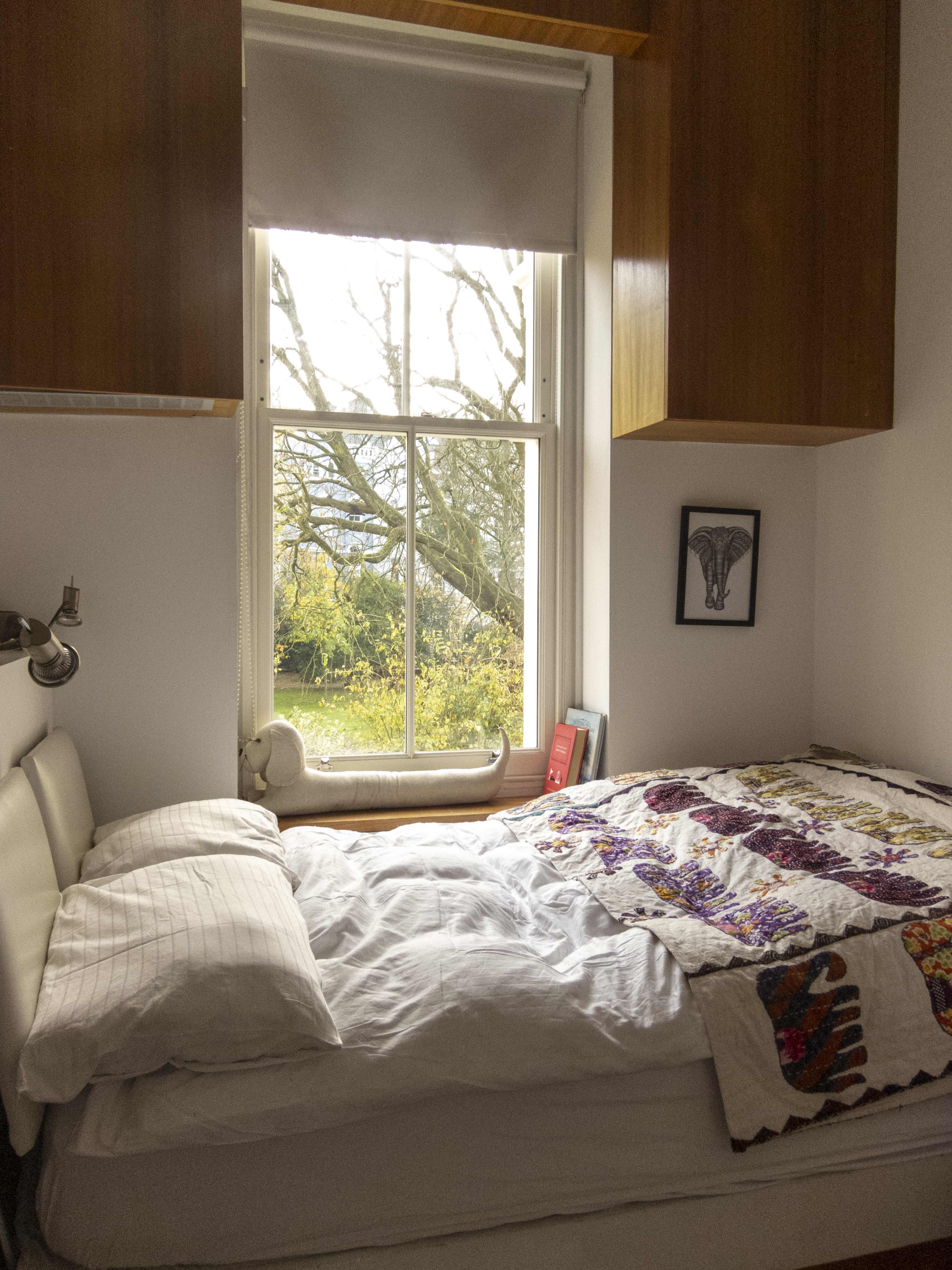 A neatly made bed is positioned beside a large window that overlooks a tree-filled garden.