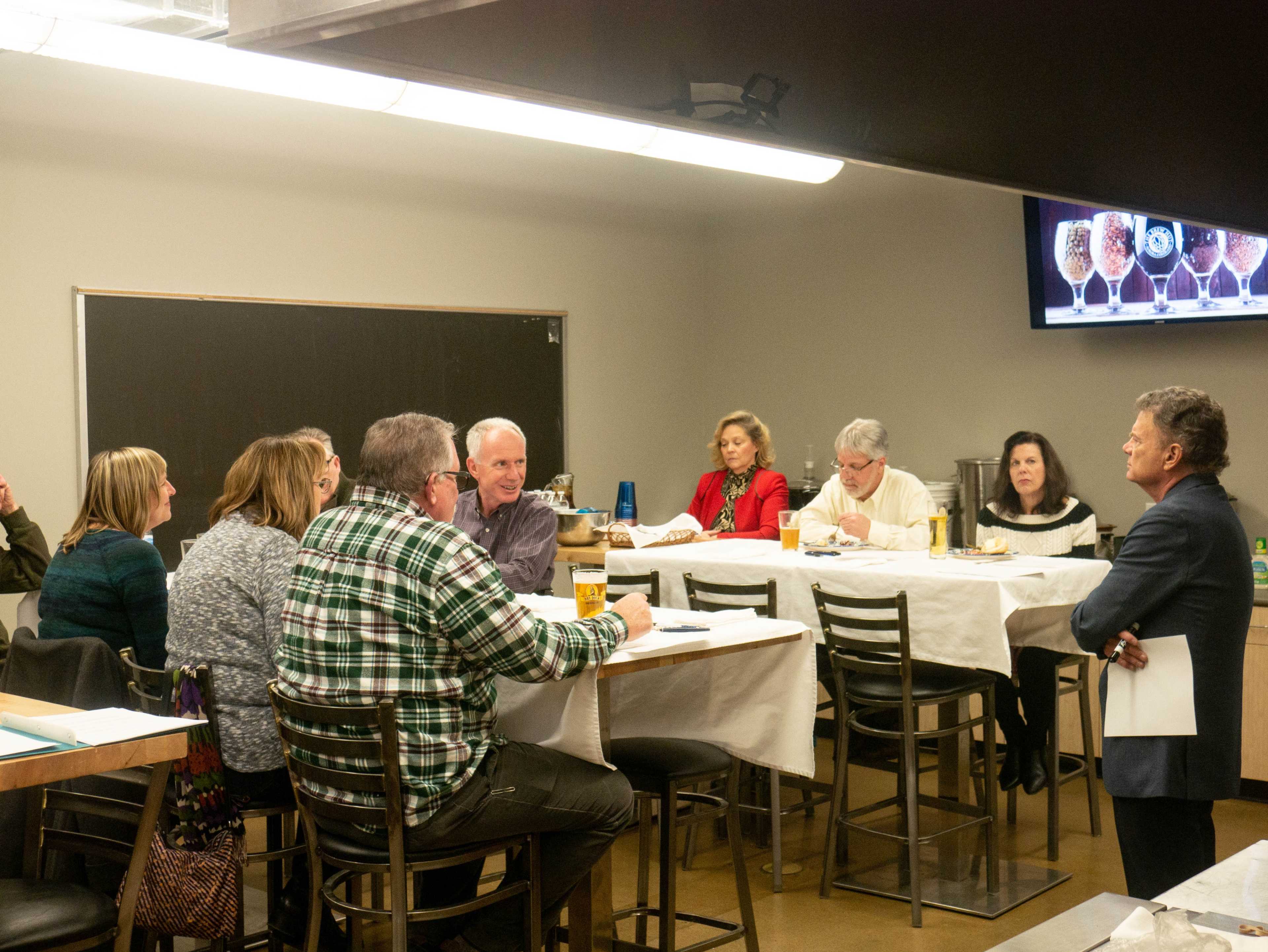 A group of people is seated around tables in a meeting room, discussing while some have papers in front of them.