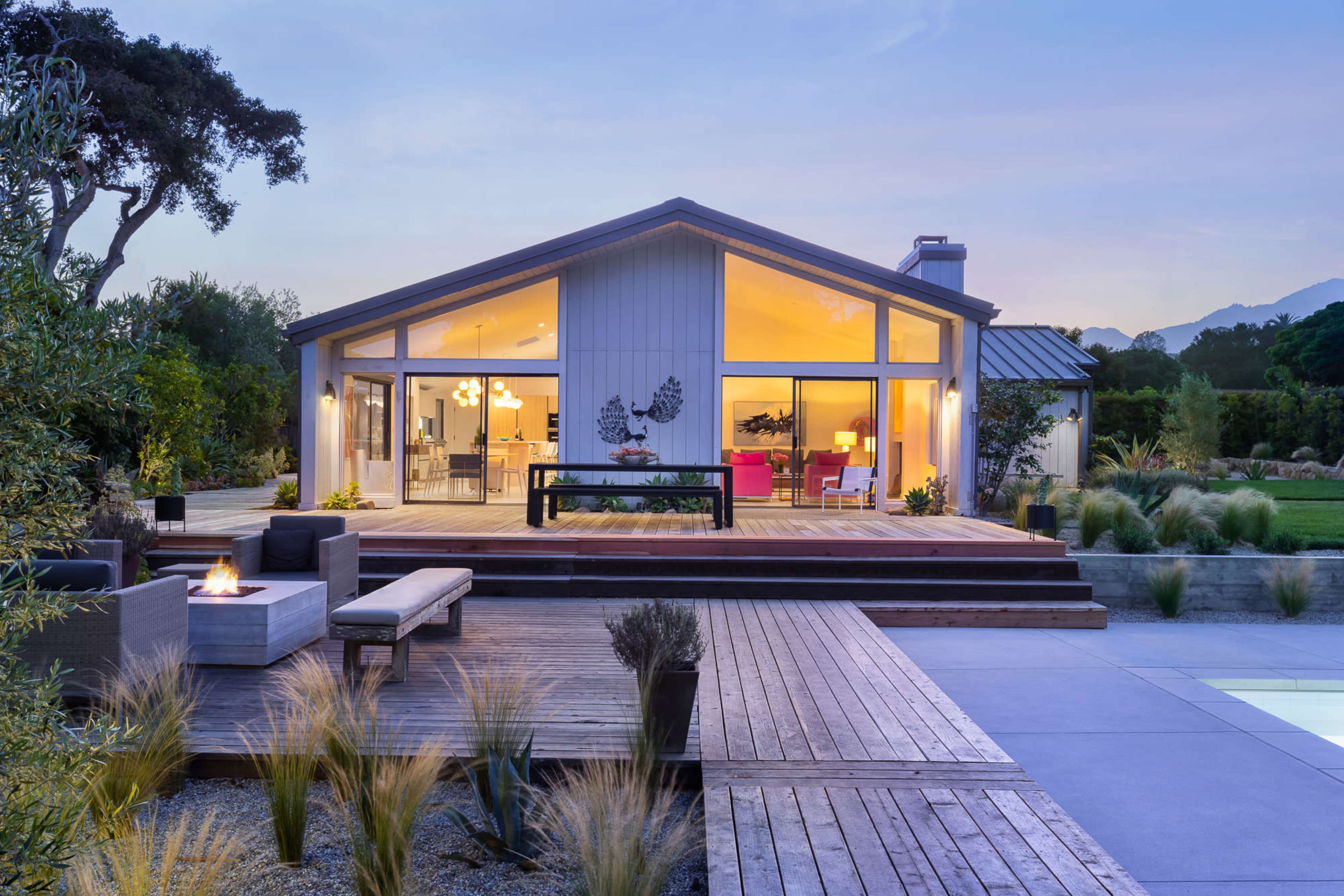A modern, single-story house with large windows and a wooden deck features a fire pit and landscaped surroundings, set against a twilight sky.
