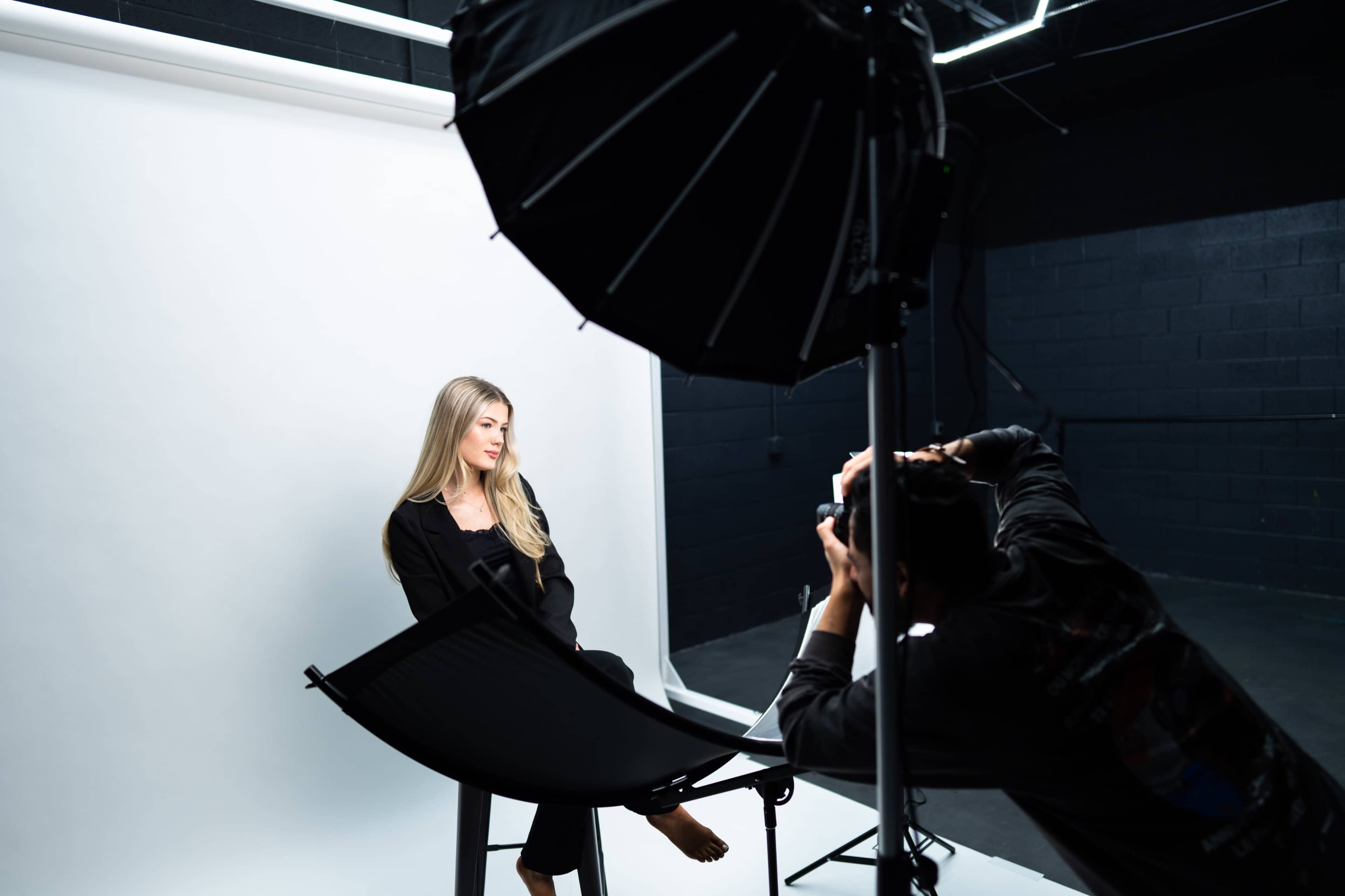 A photographer captures a model seated in a studio with a plain backdrop and professional lighting setup.