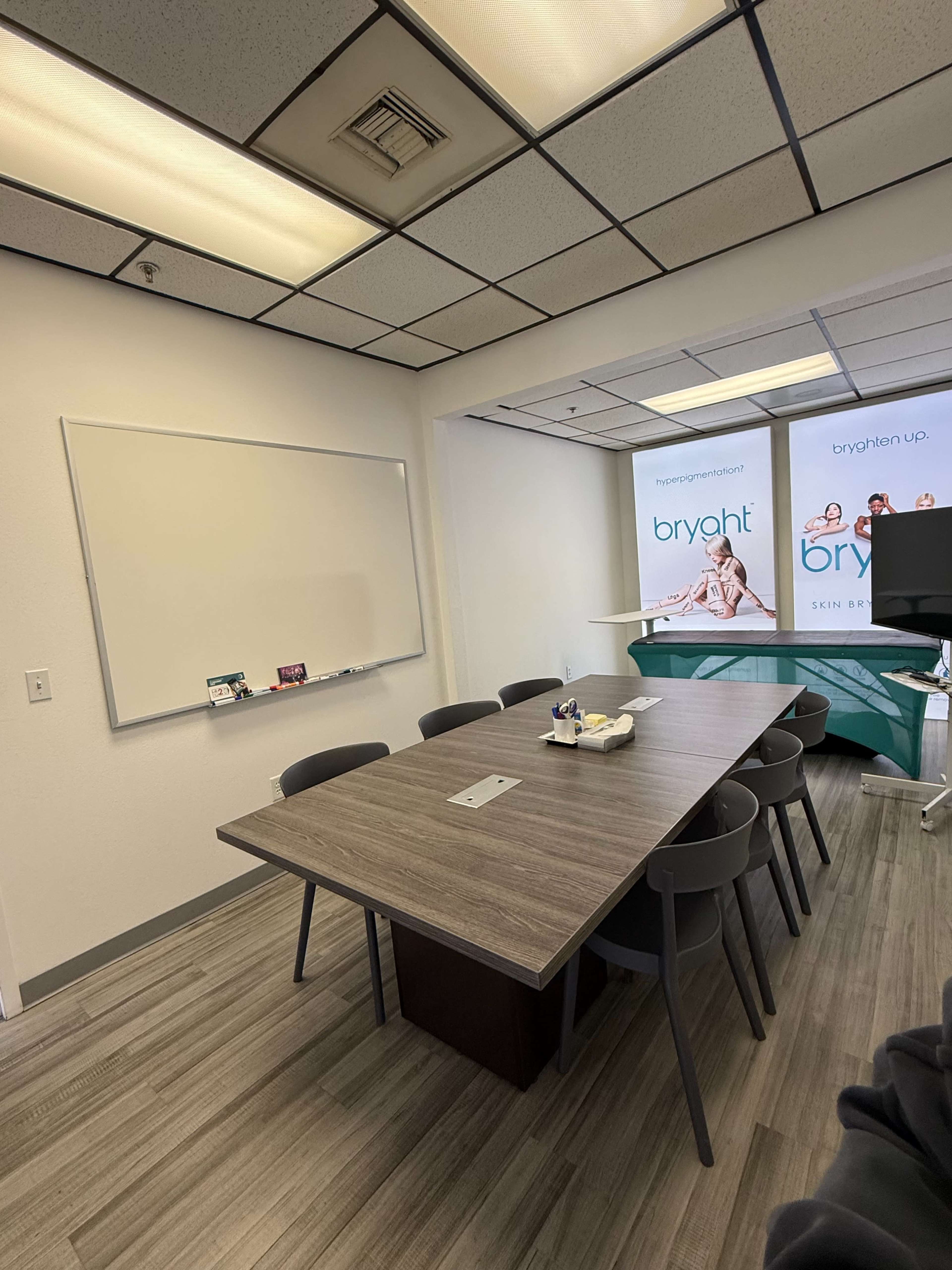 The image shows a meeting room with a long wooden table, several chairs, a whiteboard, and a display wall featuring a promotional banner.