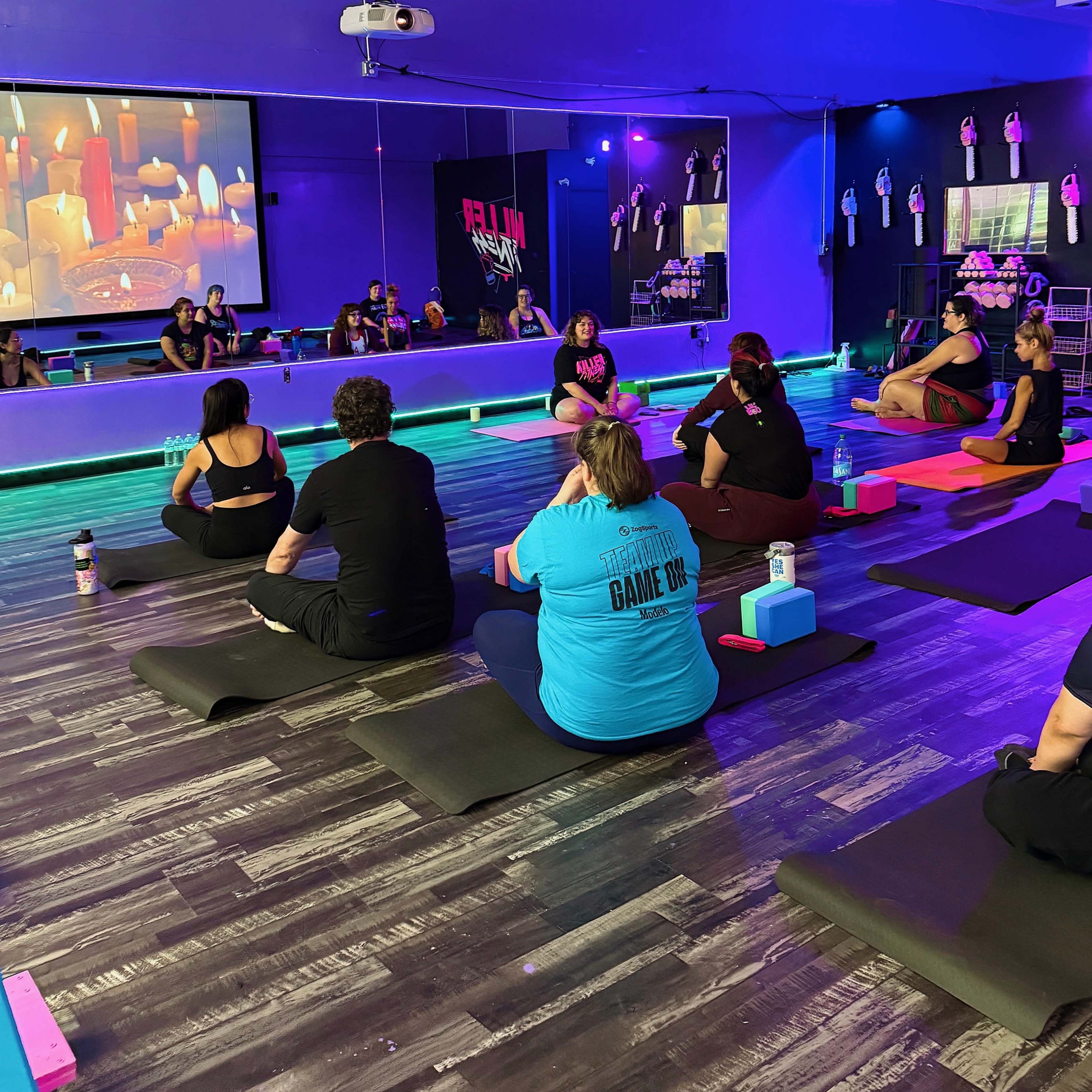 A group of people sits on yoga mats in a studio with colorful lighting and a large screen displaying images.