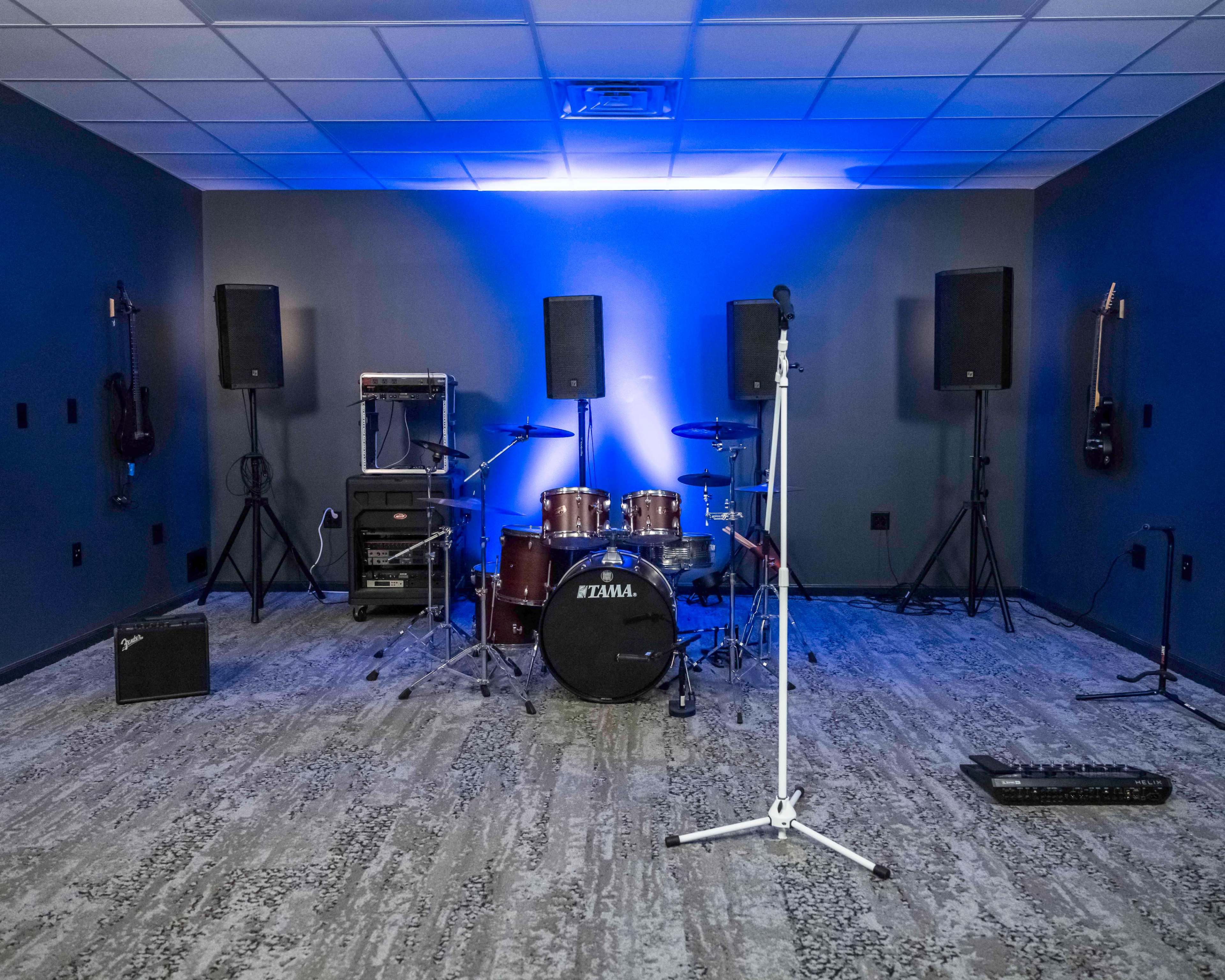 A music rehearsal space featuring a drum set, speakers, and a microphone stand against blue lighting.