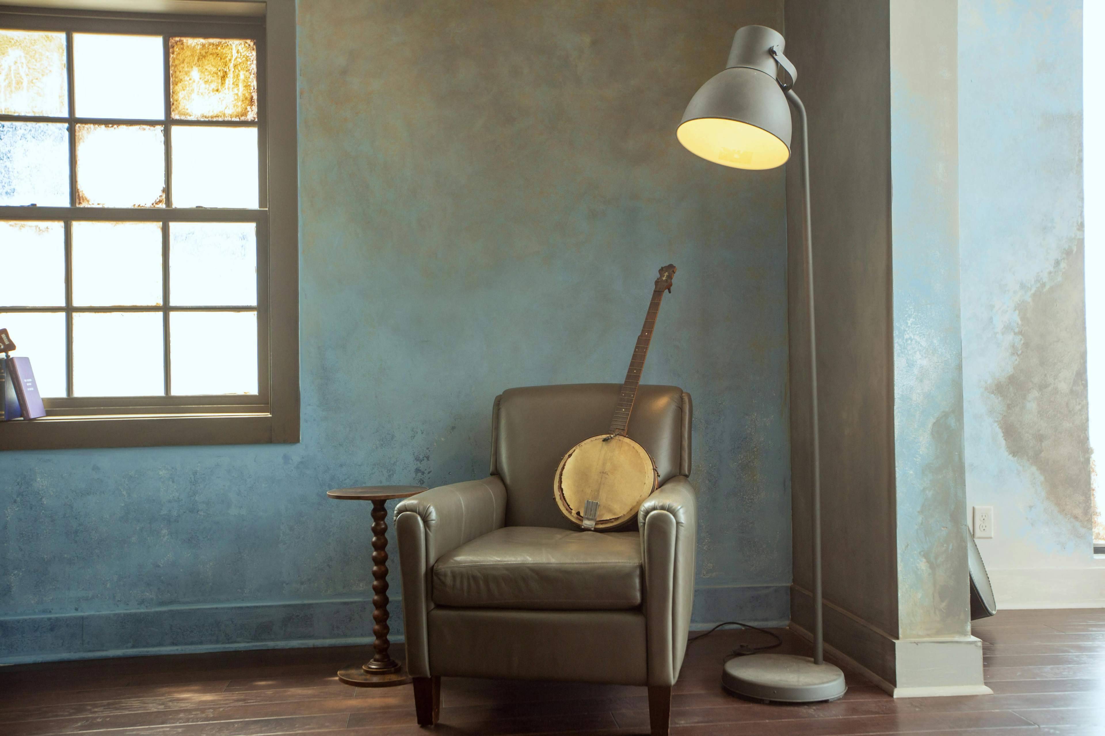A leather armchair sits beside a wooden table and a floor lamp in a room with a blue wall and a large window.