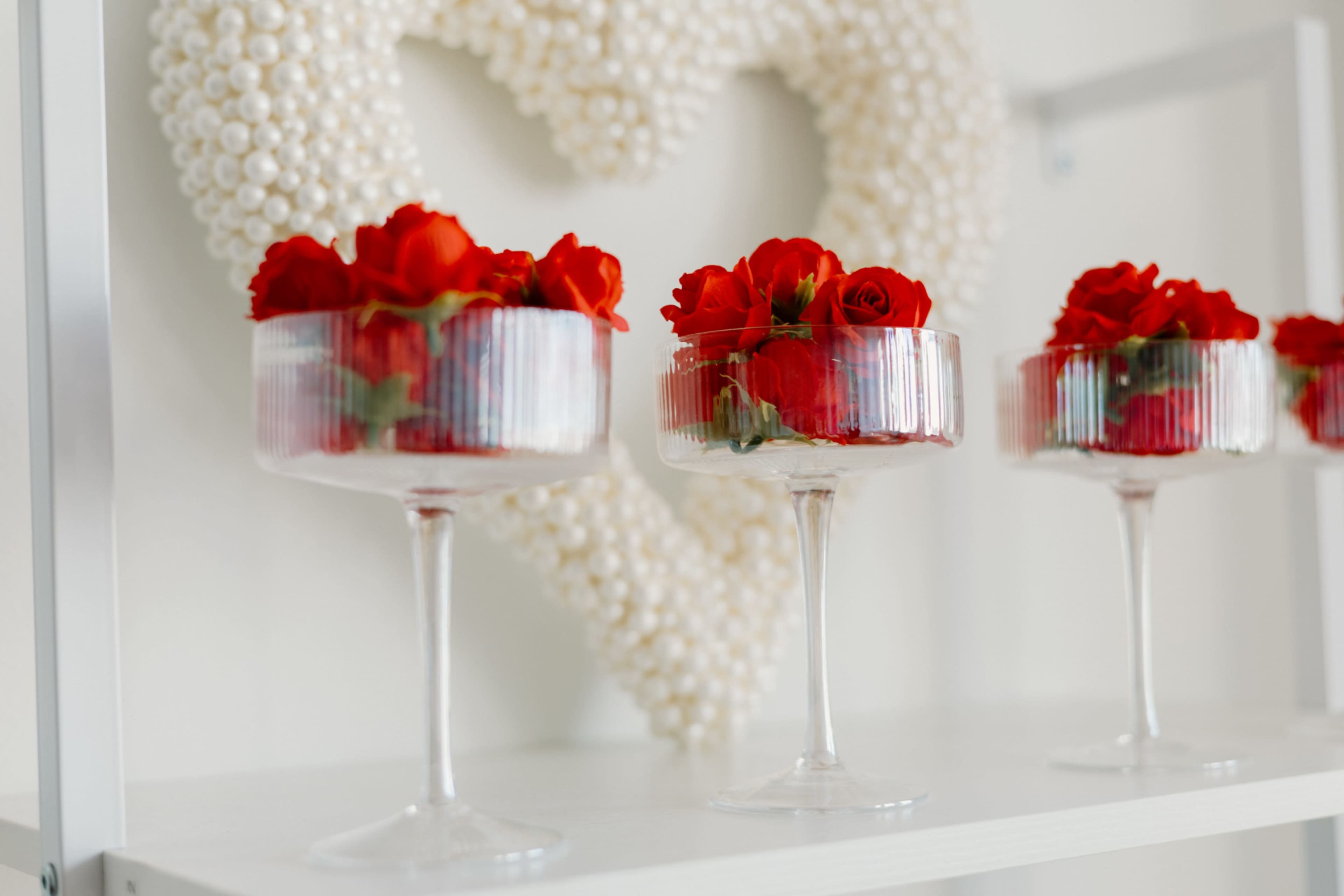 Three transparent dessert cups filled with red roses are arranged on a white shelf beneath a heart made of white pearls.