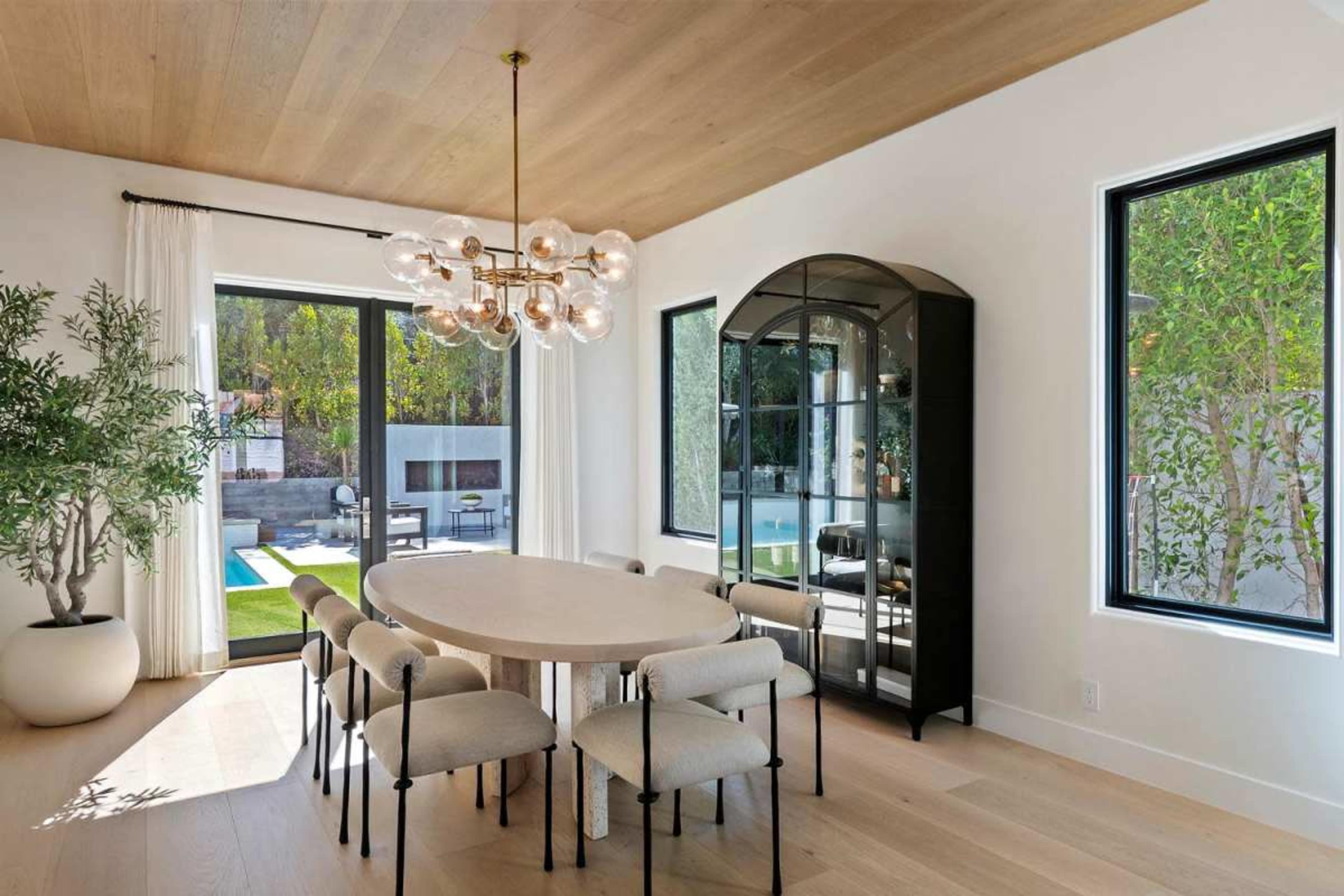 A dining room features a round table surrounded by upholstered chairs, a modern chandelier above, and large windows with views of a poolside area.
