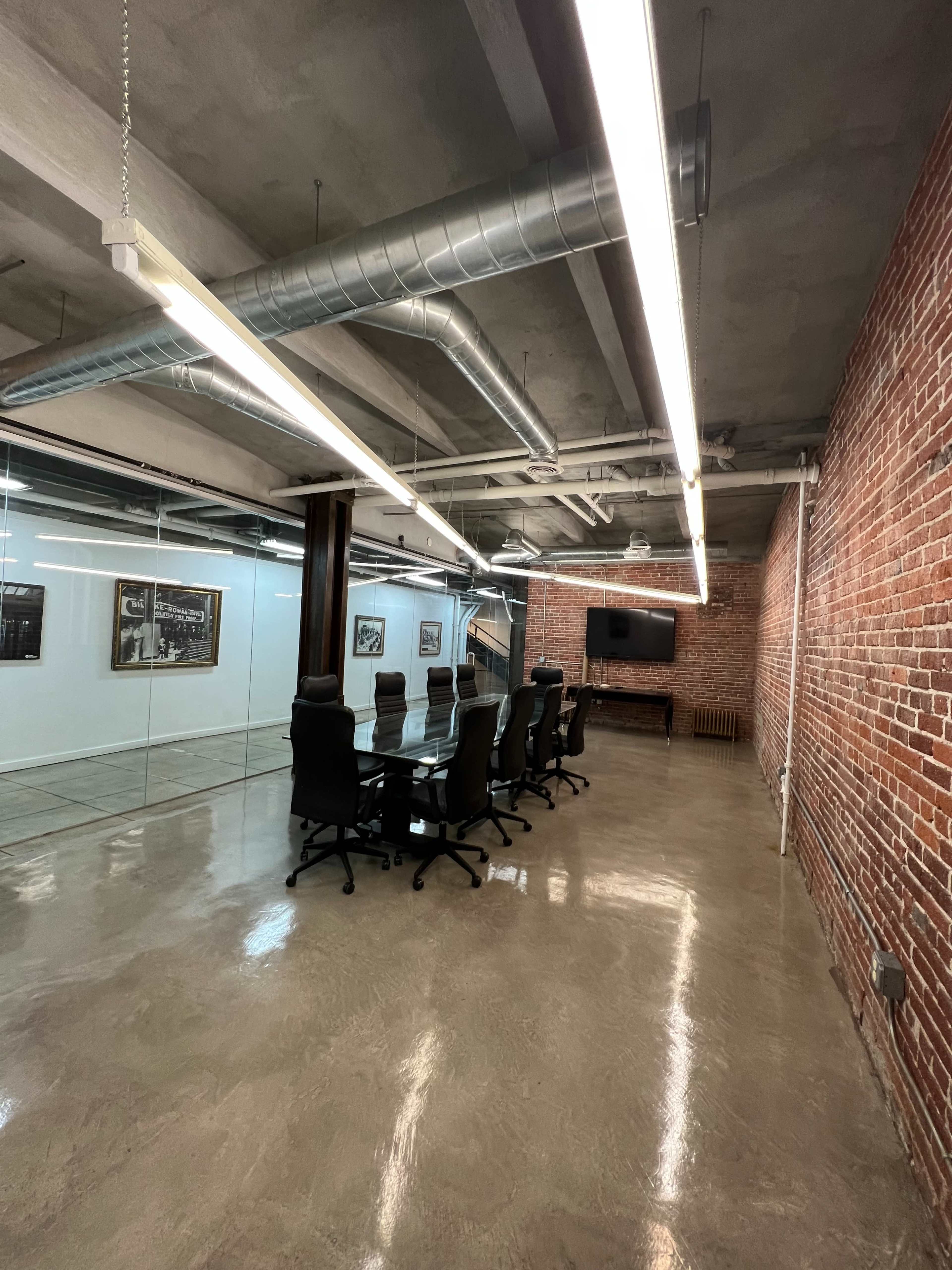 A modern conference room features a long table surrounded by black chairs, with exposed brick walls and industrial lighting.