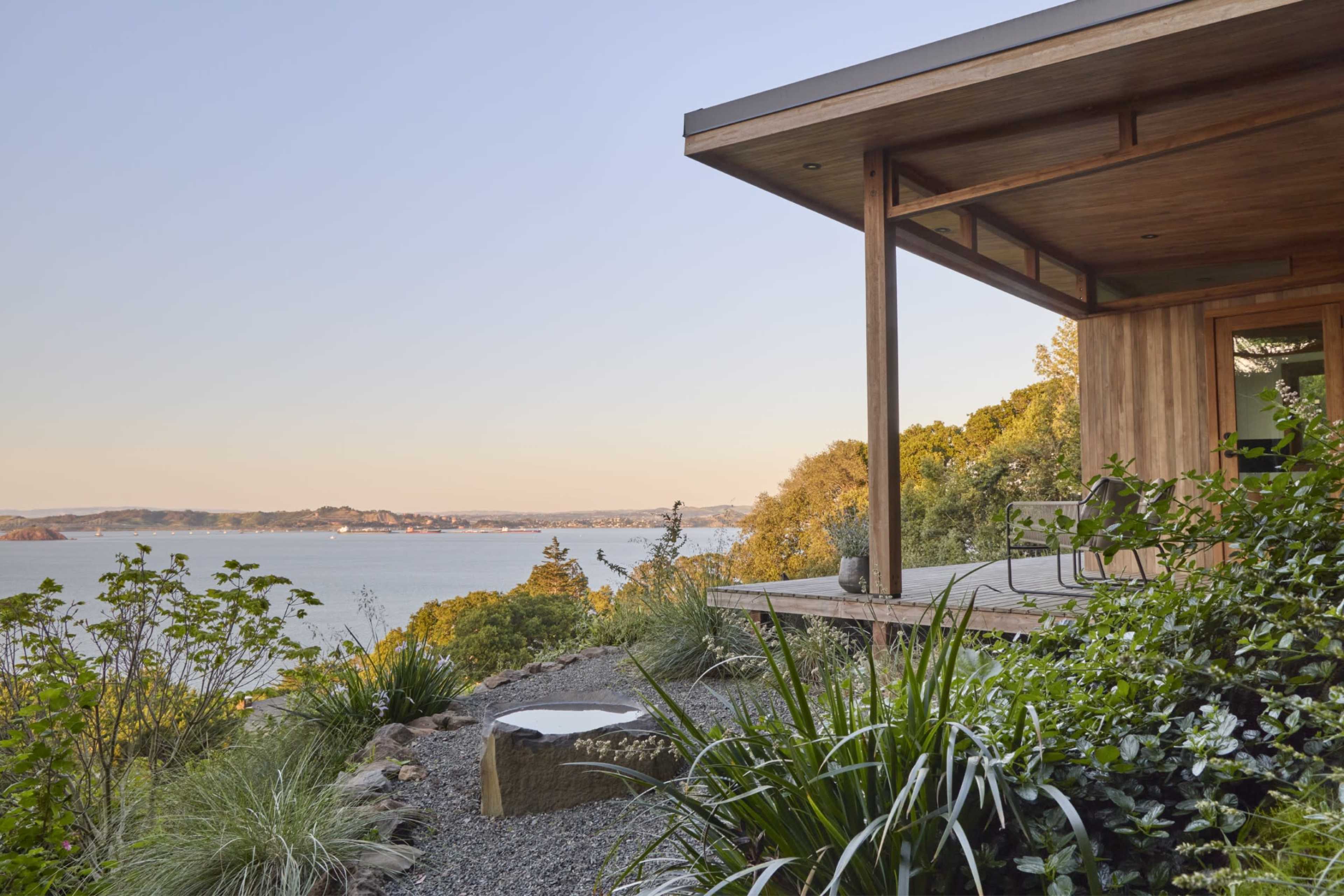 The image shows a modern wooden house with a deck overlooking a body of water and surrounding greenery.