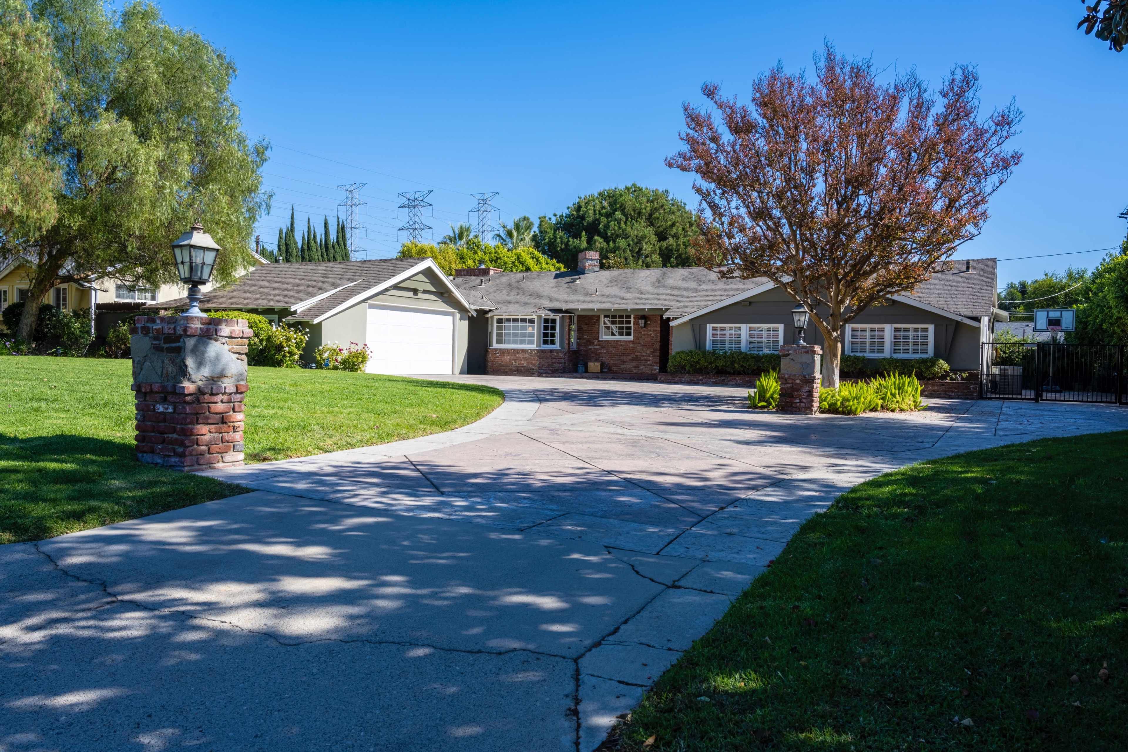 A single-story suburban house with a front lawn, a wide driveway, and a tree with red leaves.