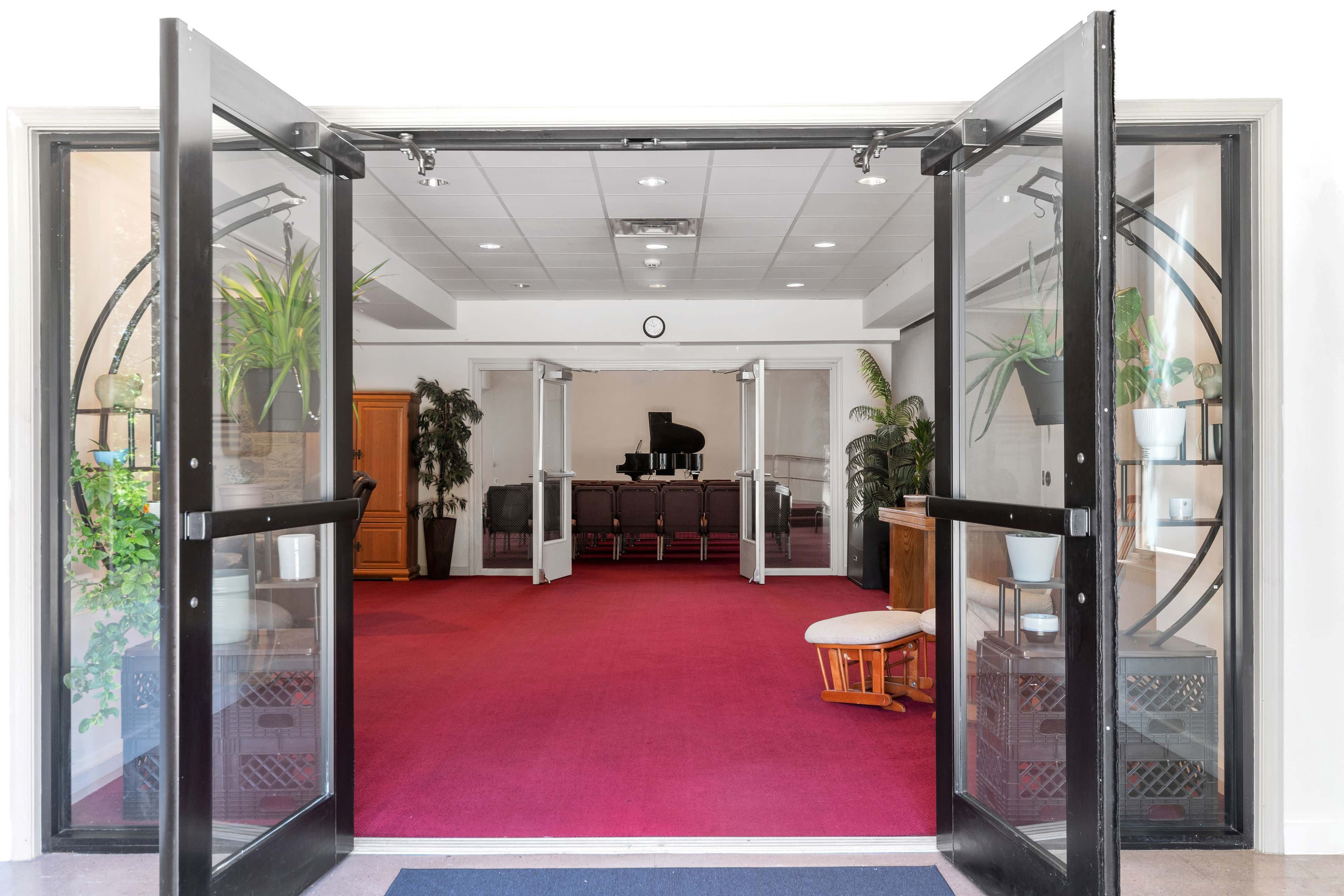 The image shows the entrance to a spacious lobby with red carpeting, featuring glass doors that open to a room containing a piano and greenery.